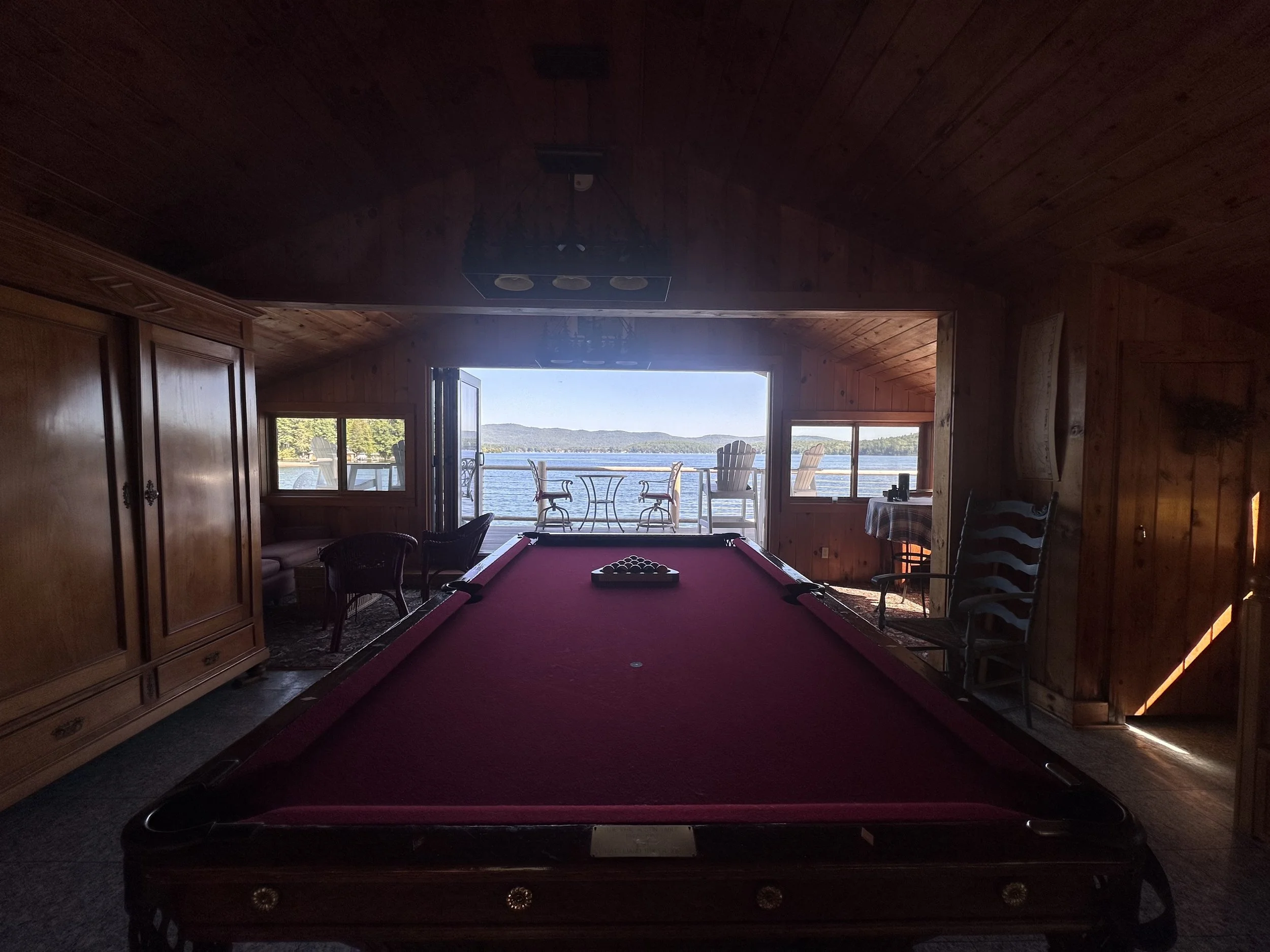 View of a lake seen from a cozy, wood-paneled room with large windows and a balcony, featuring a red pool table in the foreground and outdoor chairs on the balcony.