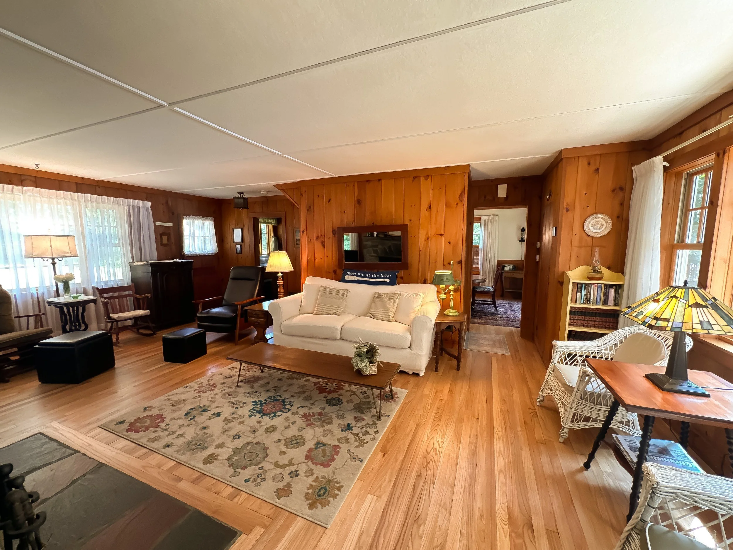 Cozy living room with wooden walls and floors, filled with vintage furniture including a white sofa, wicker chairs, and various lamps, with a rug and windows letting in natural light.