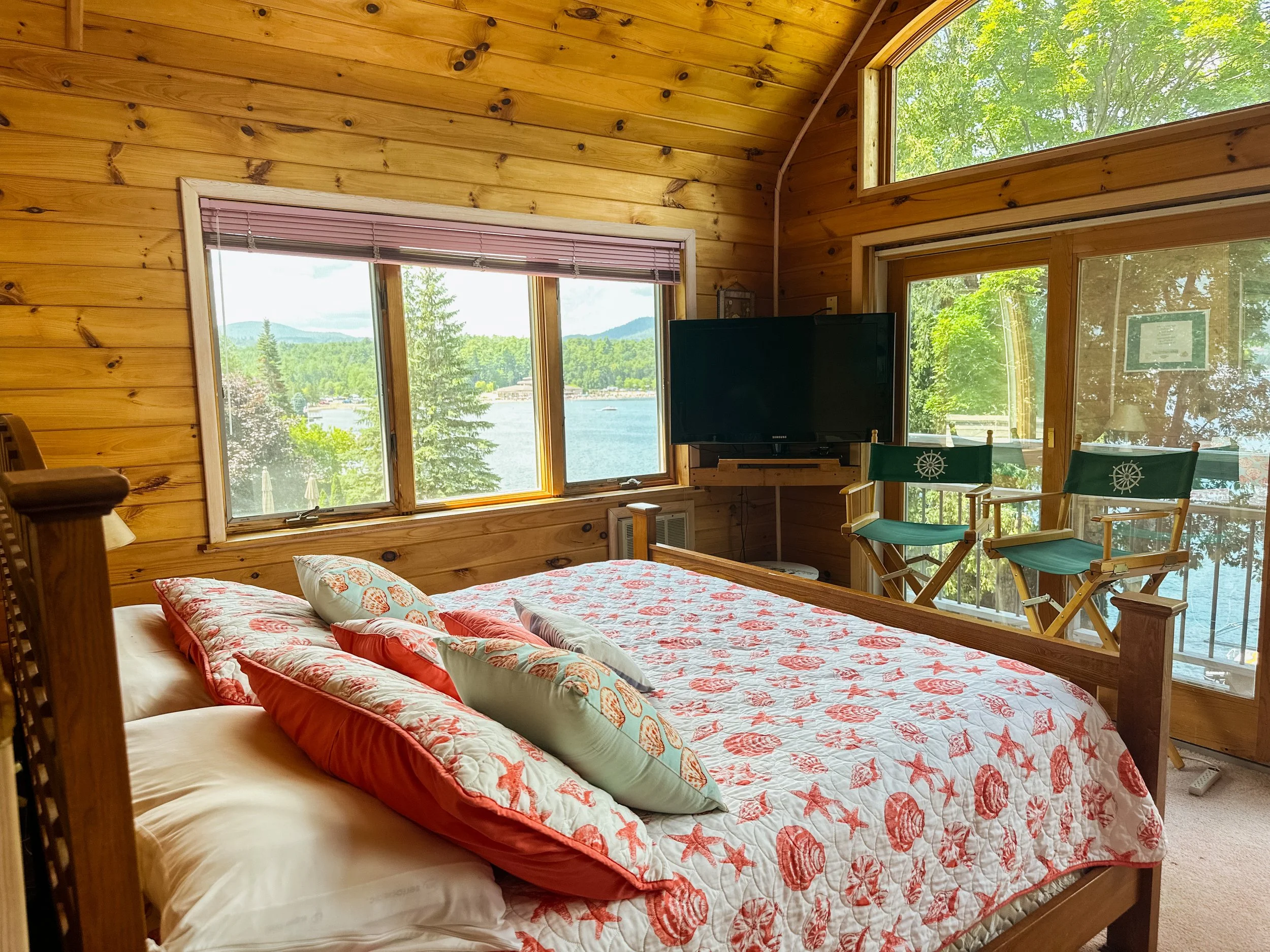 A bedroom with wood-paneled walls and large windows showing a lake and trees outside. There are two chairs with nautical steering wheel designs on the backrests, a bed with a starfish and seashell patterned quilt, and a television on a corner stand.