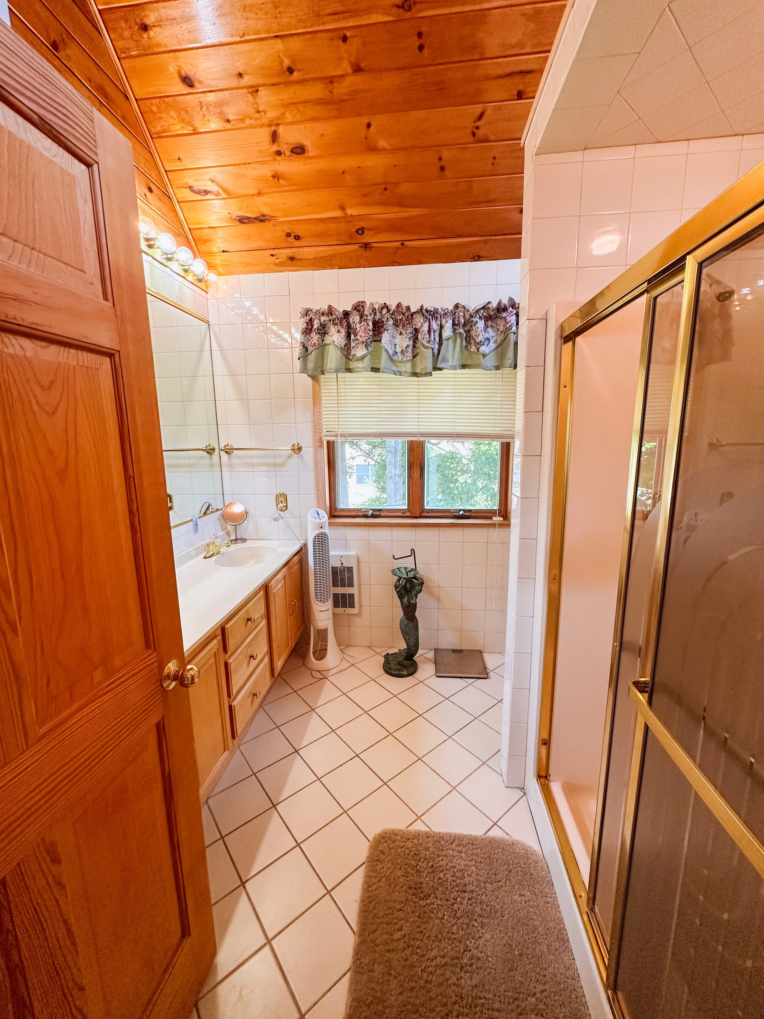 A bathroom with a wood-paneled ceiling, white tiled walls, a window with a floral curtain, and a shower with gold-colored frame on the right. There is a vanity with a sink and a mirror, and various items like a fan, a sculpture, and a scale on the fl
