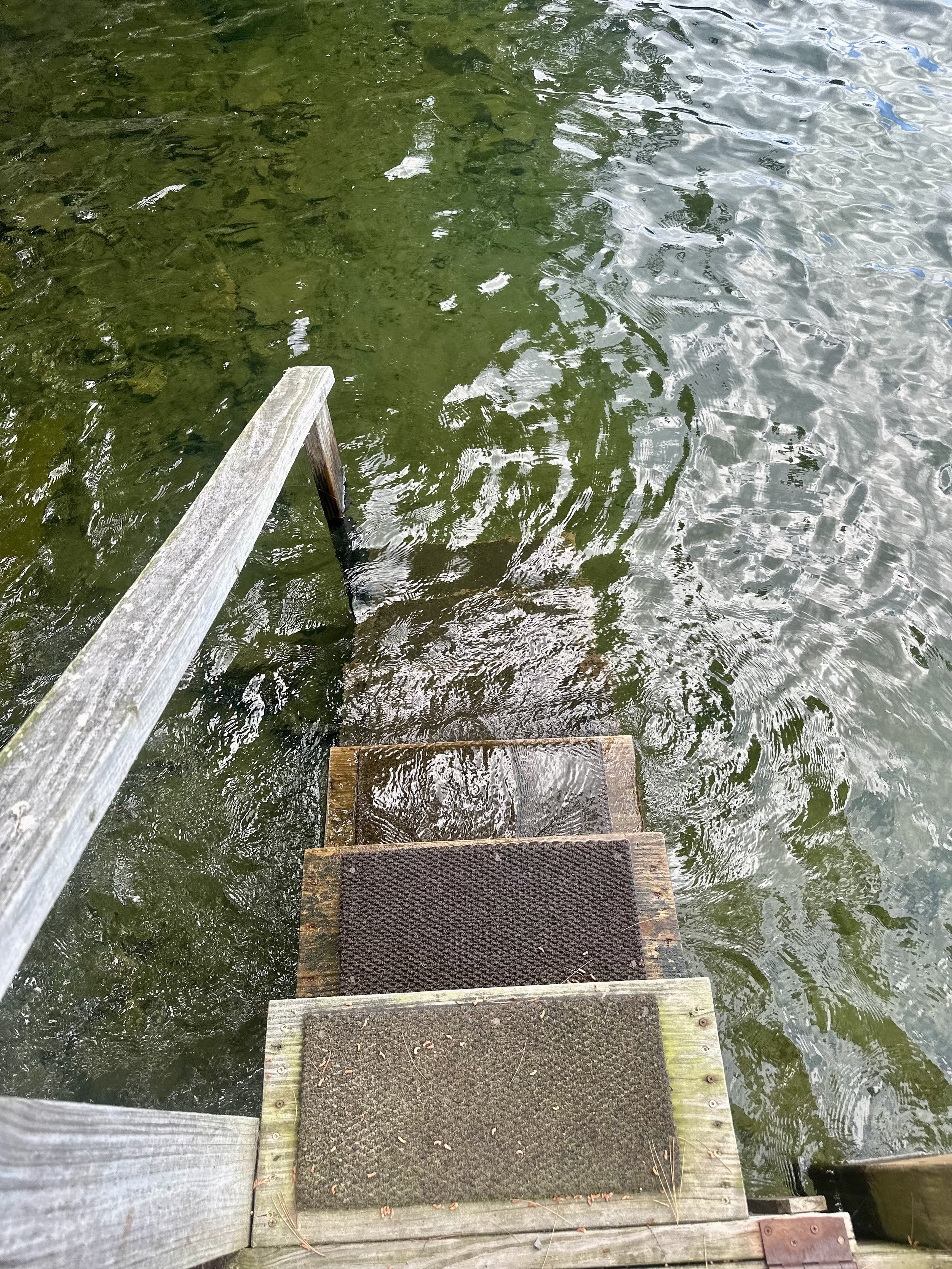 Wooden dock with steps leading into a body of water, possibly a lake or river, with reflections on the surface.