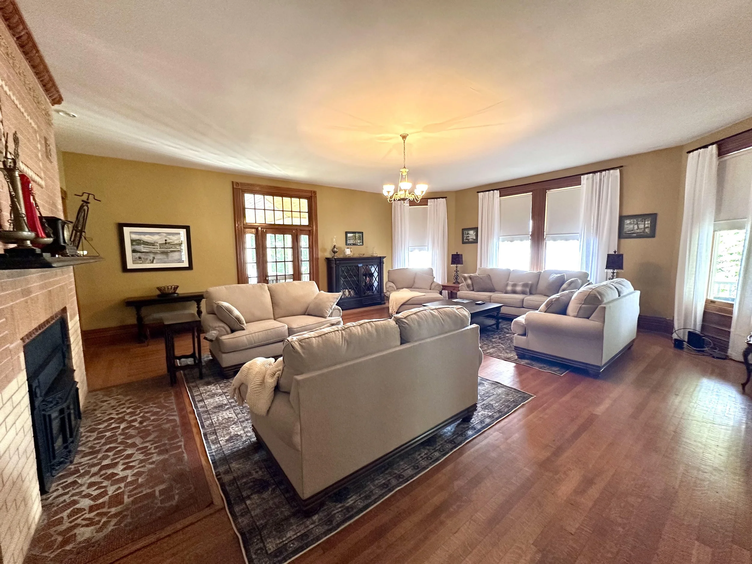 Living room with beige sofas, a wooden coffee table, a black fireplace, framed pictures on the walls, and large windows with white curtains, wooden trim, and a chandelier.