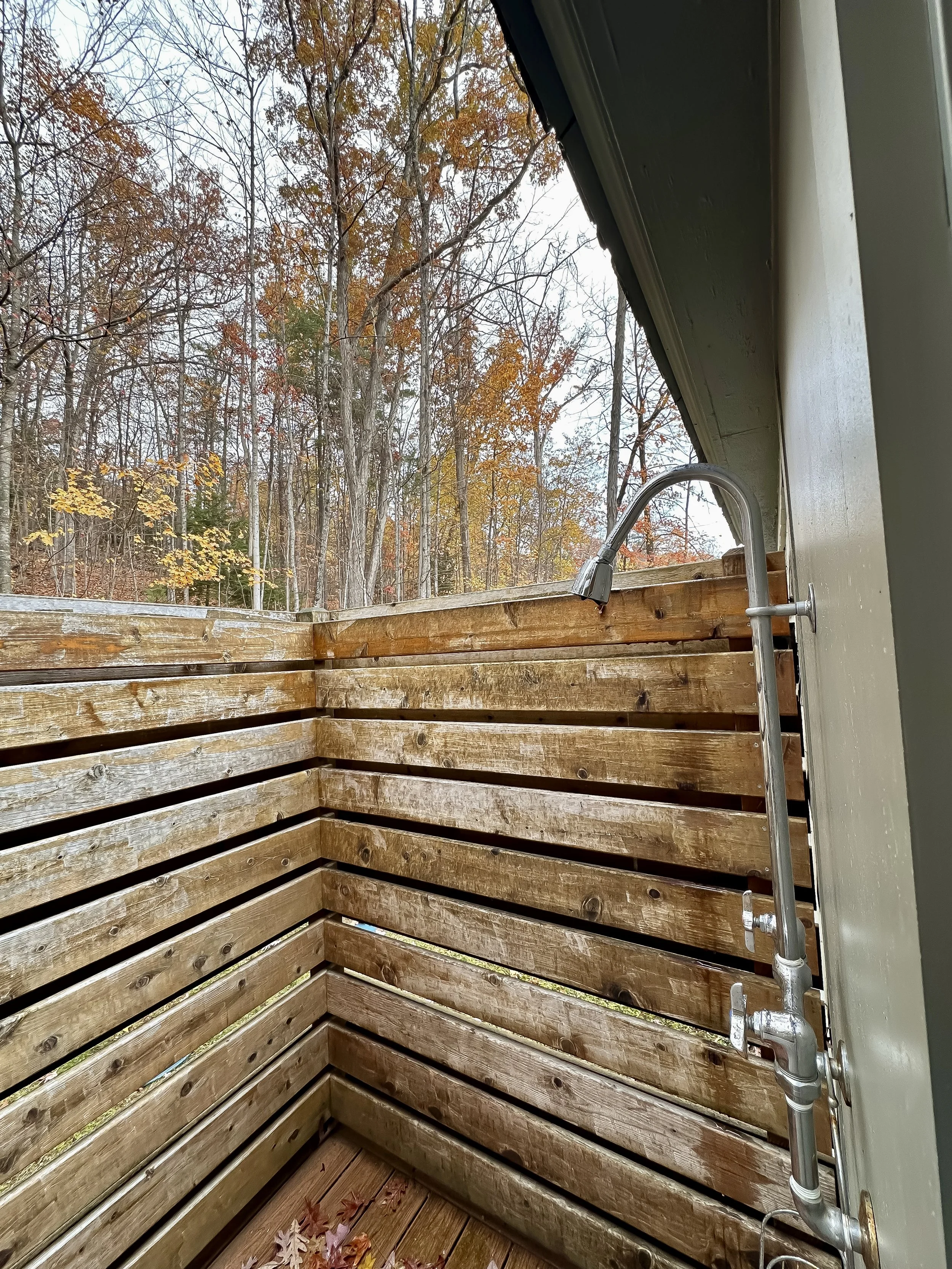 View from a small outdoor shower with a wooden privacy wall, a metal shower head, and trees with autumn foliage in the background.