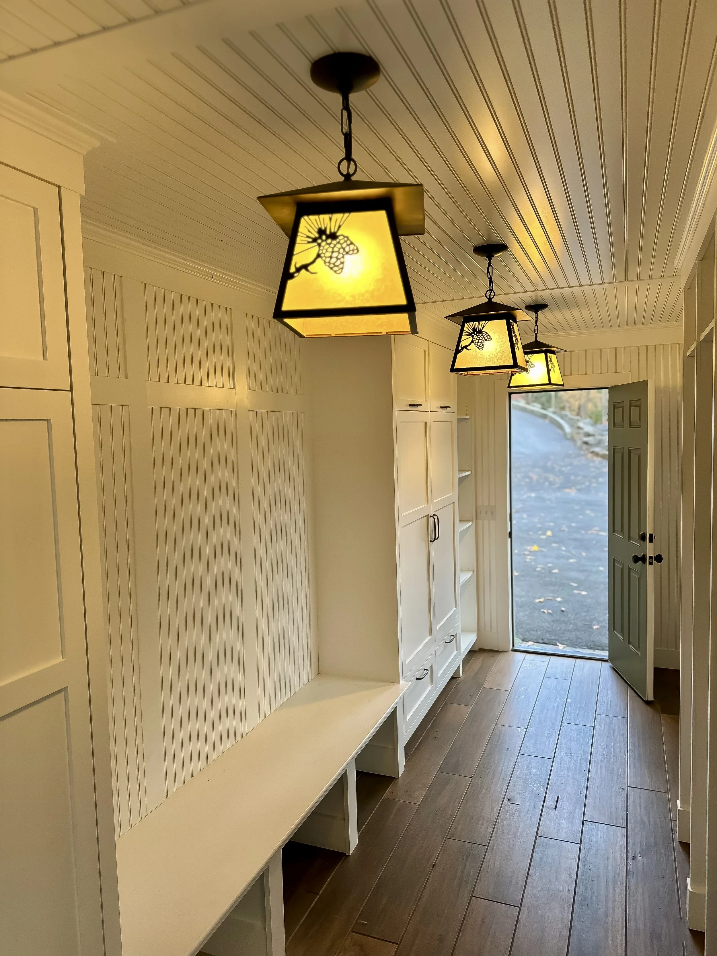 Interior of a laundry or mudroom with three hanging pendant lights with yellow shades decorated with grapevine designs, a built-in bench, white cabinets, and an open door leading outside.