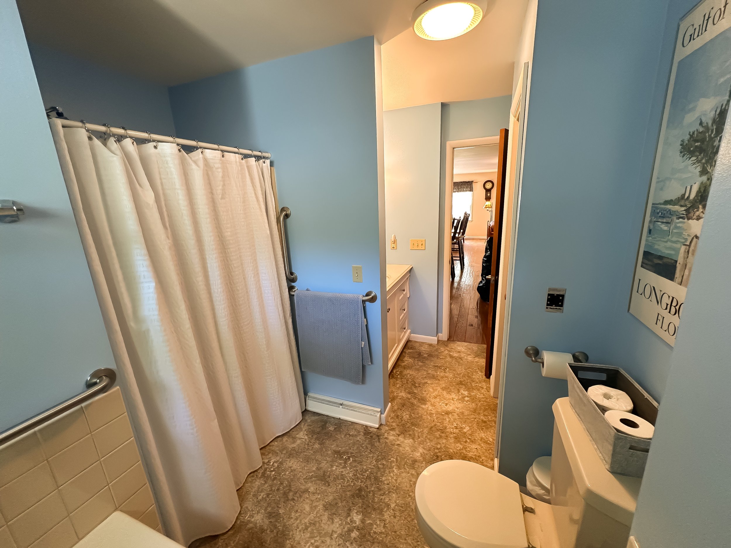 Bathroom with blue walls, a shower with a cream curtain, a toilet with a toilet paper holder nearby, a towel hanging on a bar, and a view into a dining room with a wooden table and chairs.