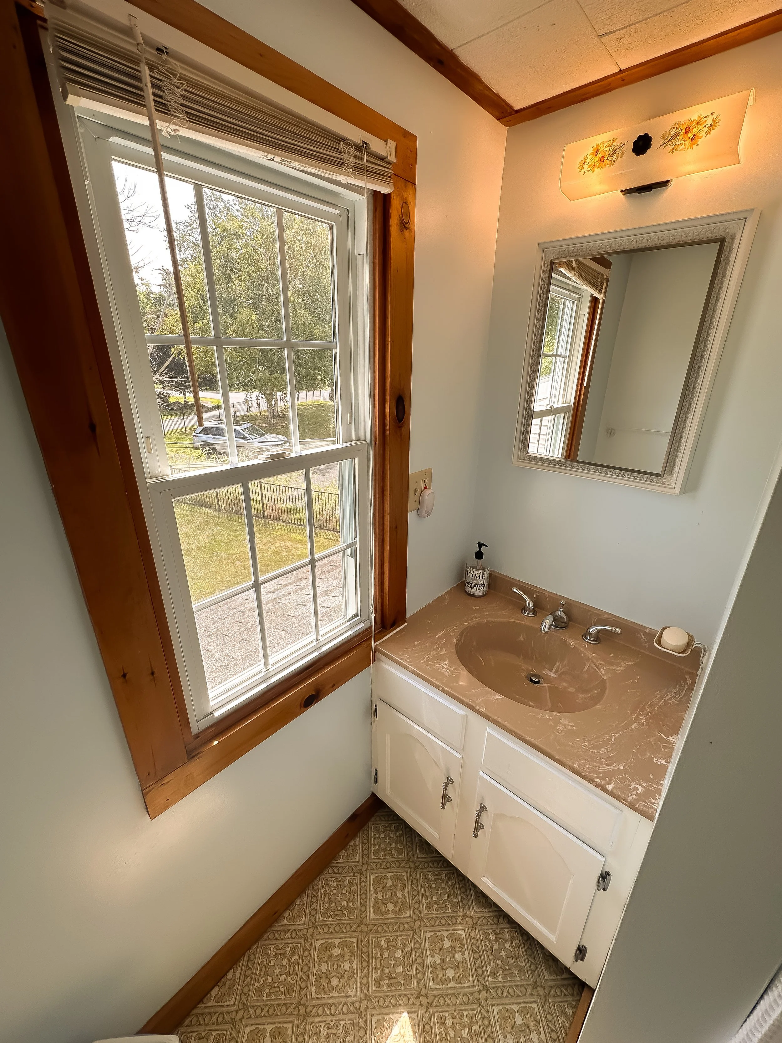 A small bathroom corner with a window, a white-framed mirror, a beige sink with a marble countertop, a soap dispenser, a soap bar, and a wall-mounted light fixture with a floral design.