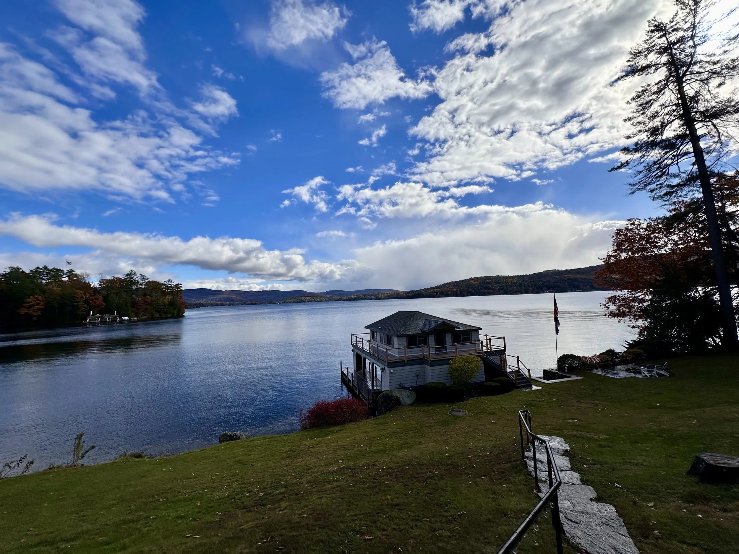 View of a lake with a house built on the water's edge, surrounded by trees with autumn foliage, under a partly cloudy sky.