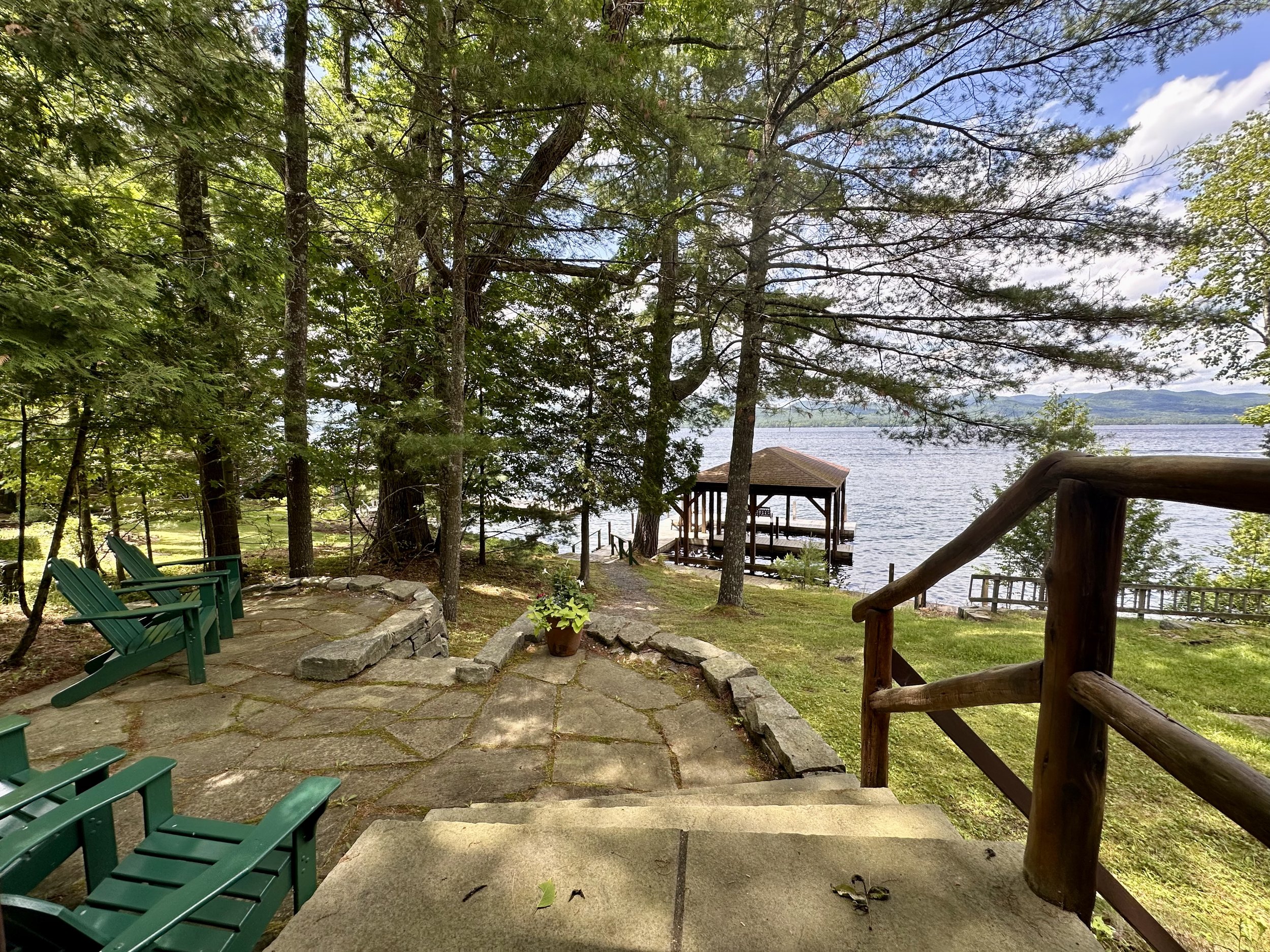 A lakeside scene with a stone patio, green chairs, a potted plant, a wooden railing, and a dock with a pavilion over calm water, surrounded by trees under a partly cloudy sky.