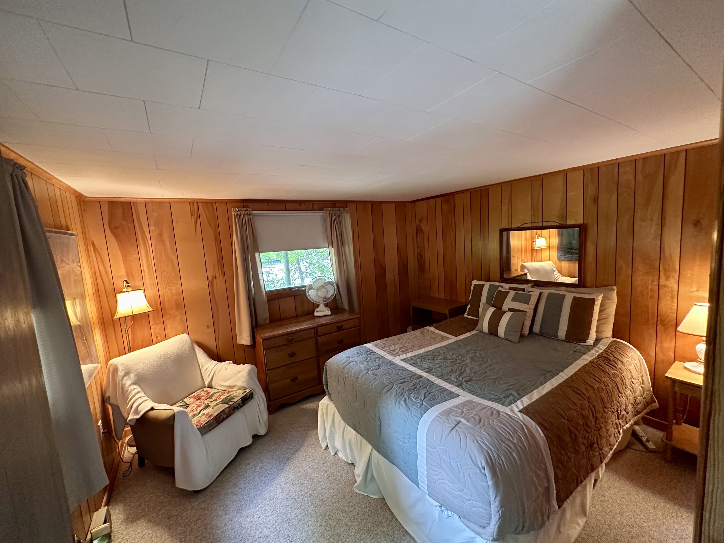 A cozy bedroom with wood-paneled walls, a bed with a gray and brown quilt and multiple pillows, a small white armchair with a throw blanket, a wooden dresser with a fan, a mirror, two bedside tables with lamps, a window with curtains, and a small pai