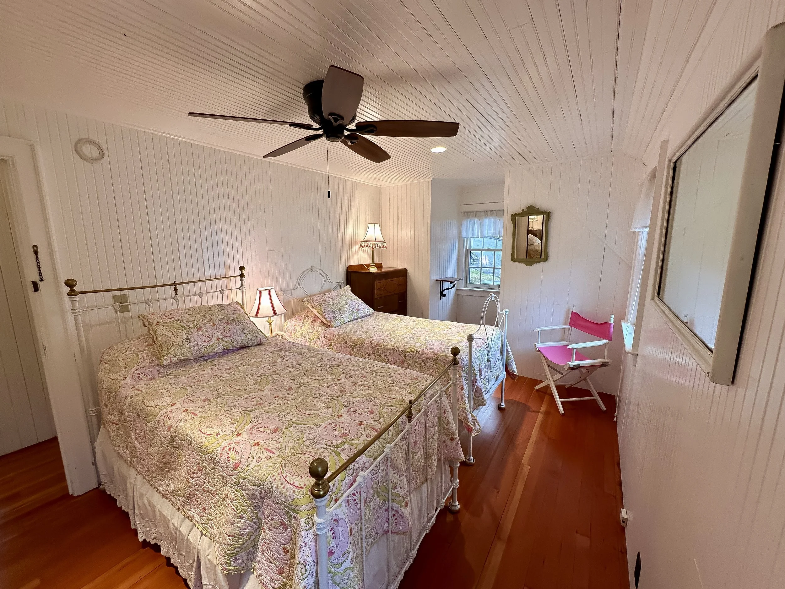 A cozy bedroom with two twin beds with floral bedspreads, a wooden dresser, two small lamps, a ceiling fan, a window with curtains, a pink folding chair, and a mirror on the wall.