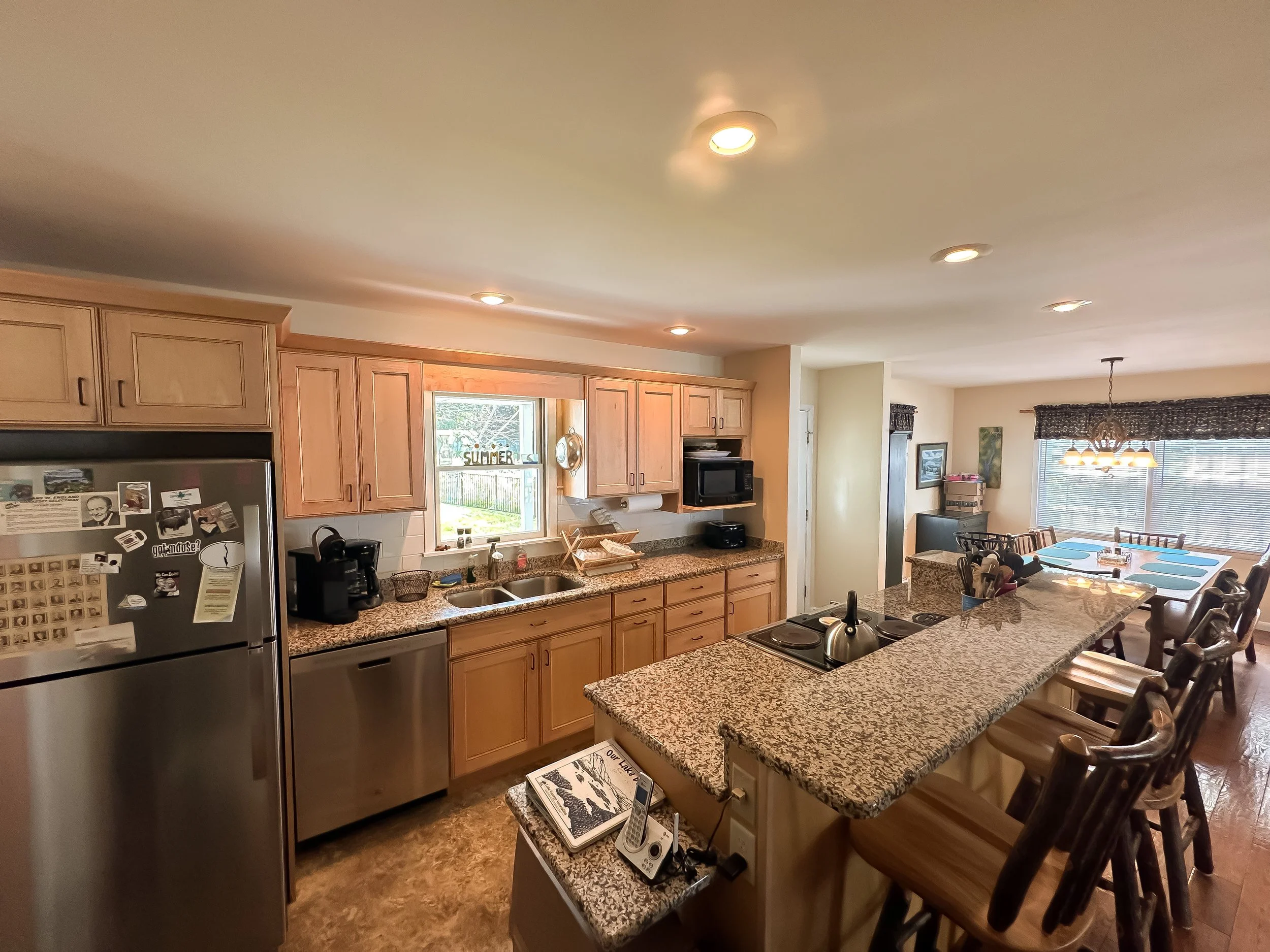 Kitchen and dining area with granite countertops, wooden cabinets, stainless steel refrigerator, microwave, coffee maker, sink under window, and a dining table with chairs. Bright, well-lit with ceiling lights and large window with blinds.