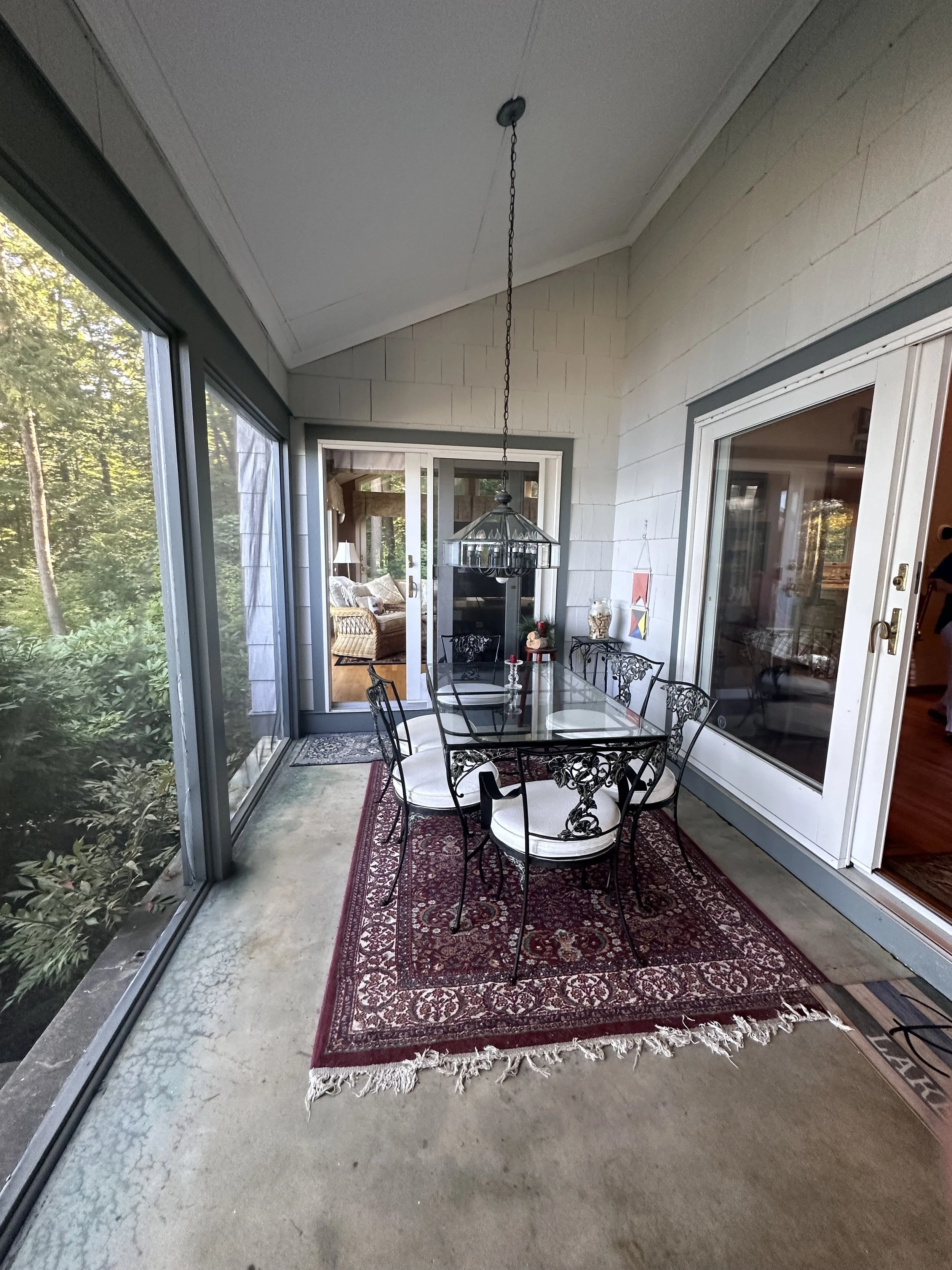 A screened porch with a glass-top dining table and six ornate metal chairs with white cushions. There is a red patterned rug under the table, a hanging lantern-style light fixture, and a view of a room with a fireplace and windows through sliding gla
