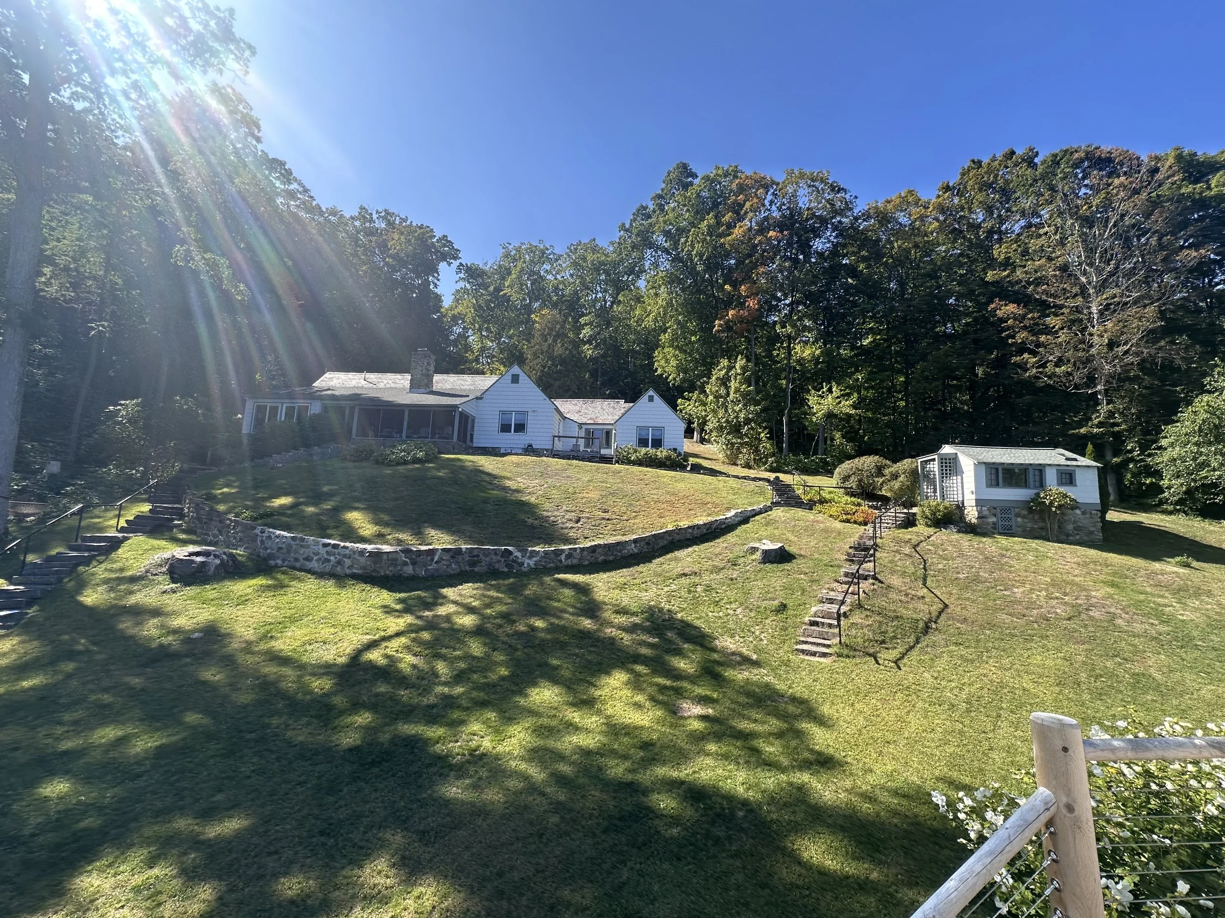 A large house on a grassy hillside with stairs and a stone wall, surrounded by trees under a clear blue sky, with sunlight creating lens flare.