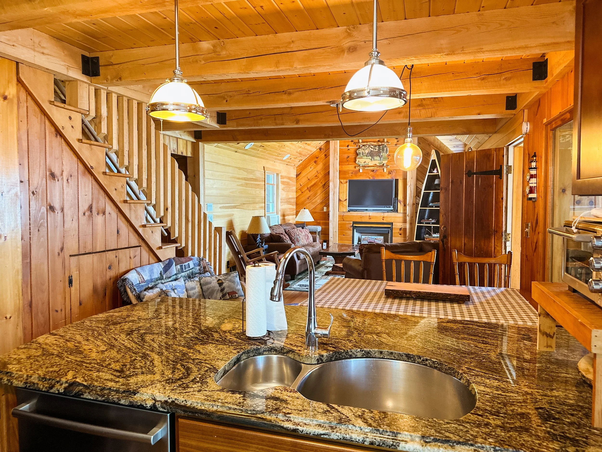Interior view of a rustic cabin with wooden walls and ceiling, featuring a stone kitchen island with a sink, modern pendant lights, and a cozy living room with a sofa and television.