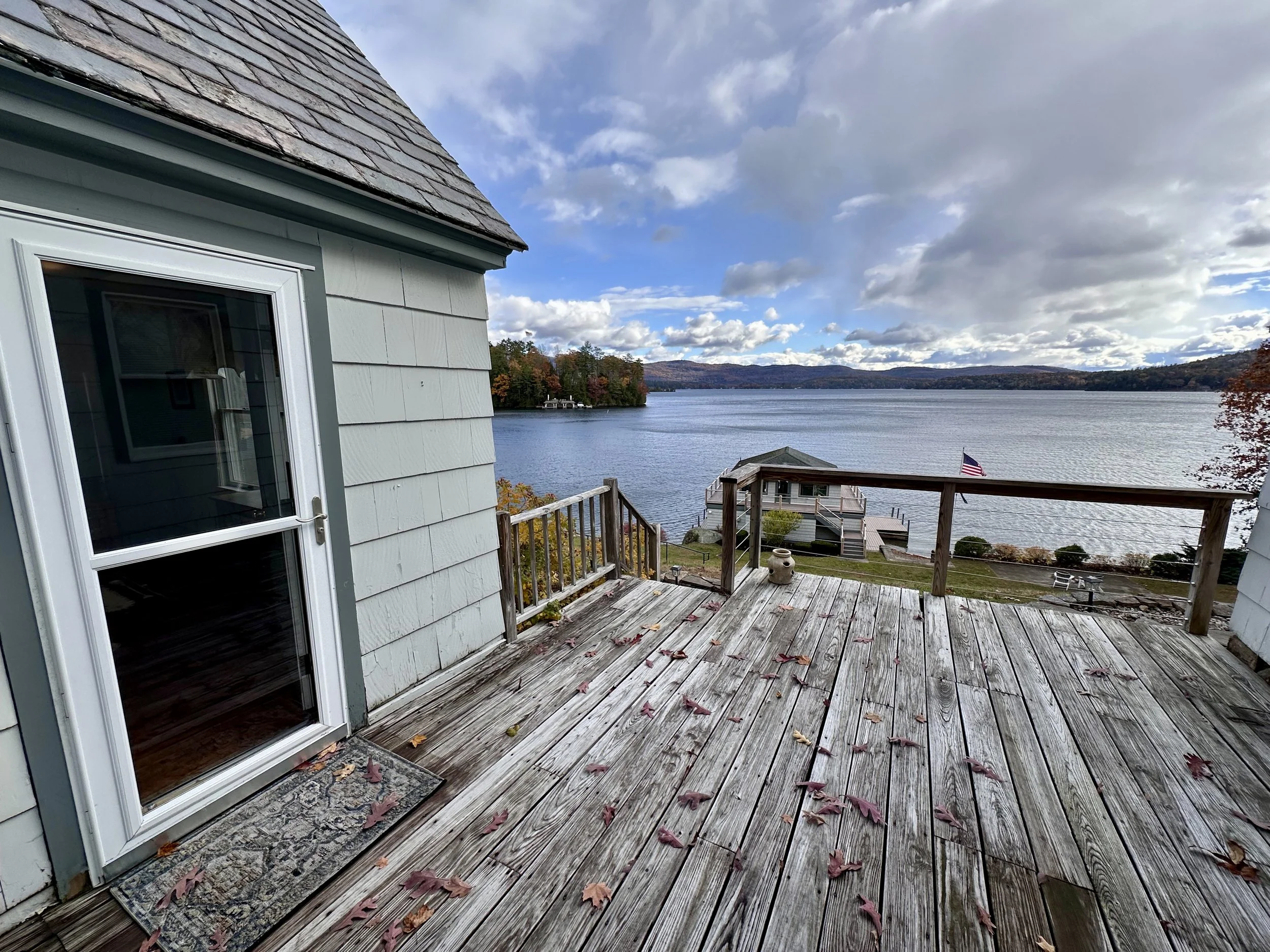 A wooden deck attached to a house overlooking a large body of water with trees displaying fall foliage. The sky has clouds with some patches of blue, and an American flag is on a boat near the shore.