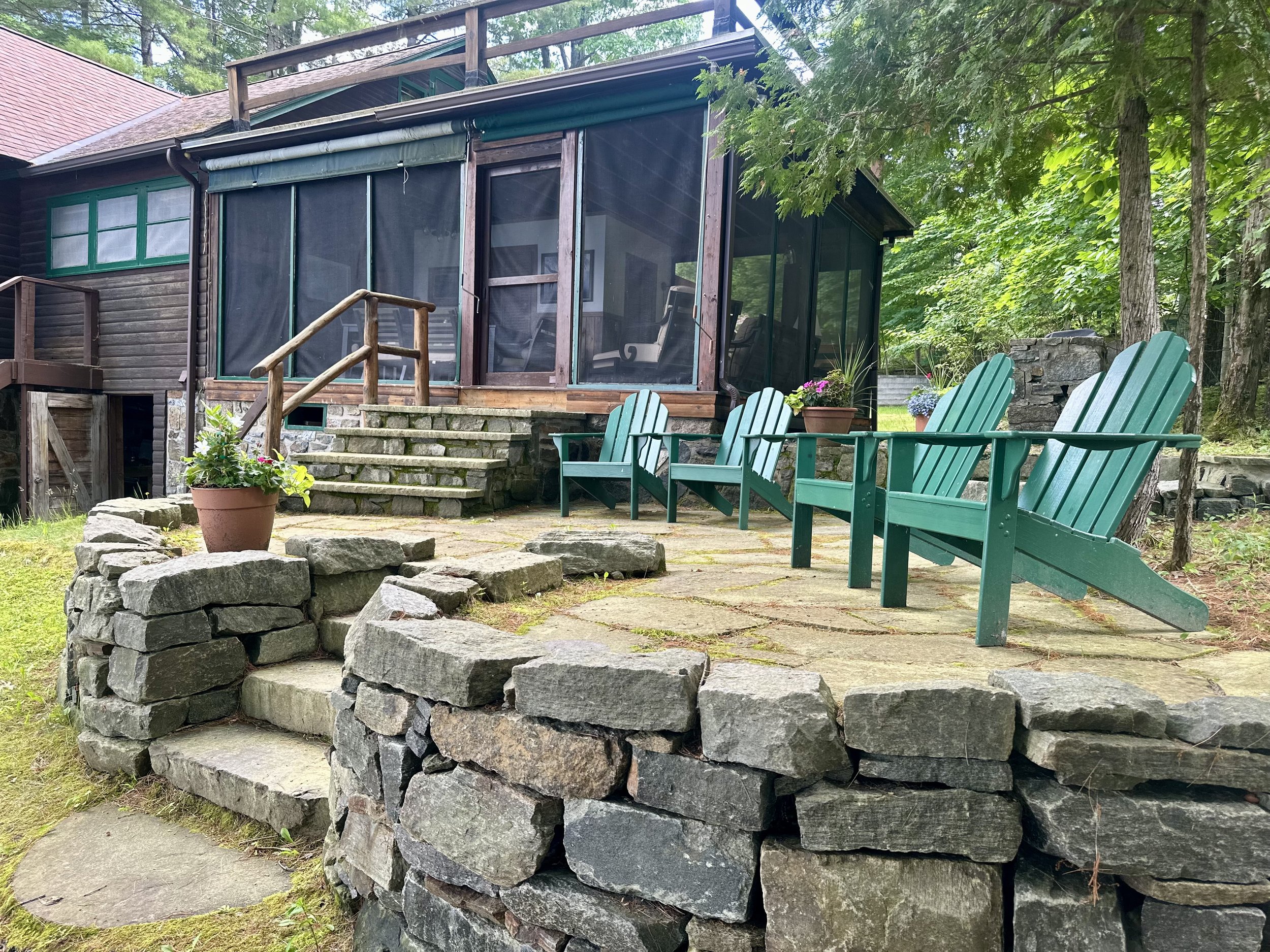 A house with a screened porch, green Adirondack chairs on a stone patio, surrounded by trees and landscaping.