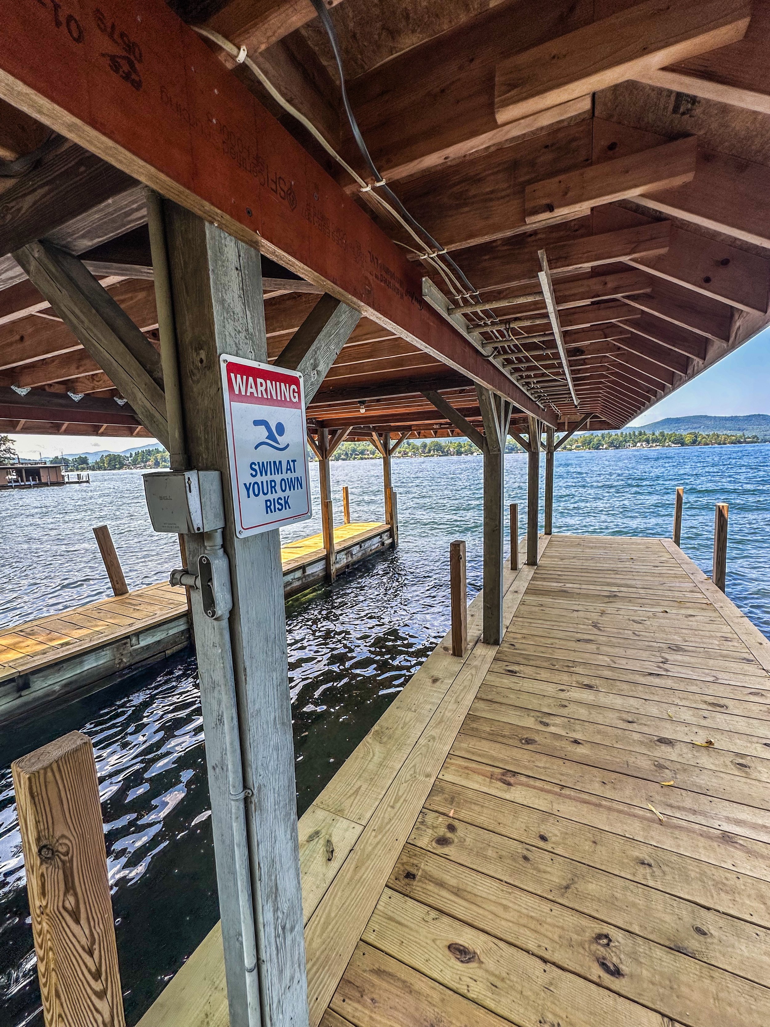 Wooden dock extending over water with a covered pavilion, and a warning sign that says 'Swim at Your Own Risk'.