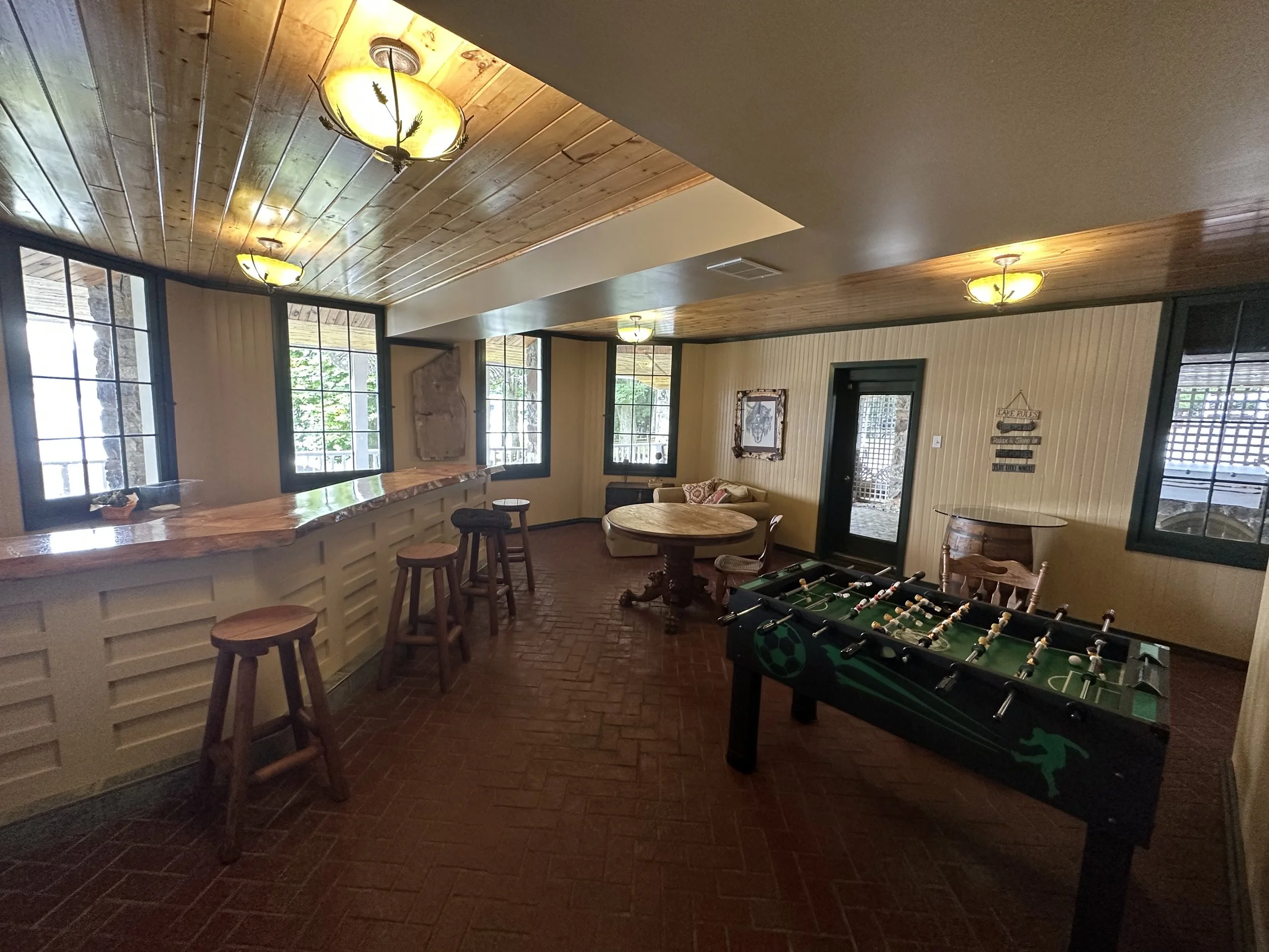 An indoor room with wooden ceiling panels and large windows, furnished with a round table, bar stools, a foosball table, and a cozy seating area.