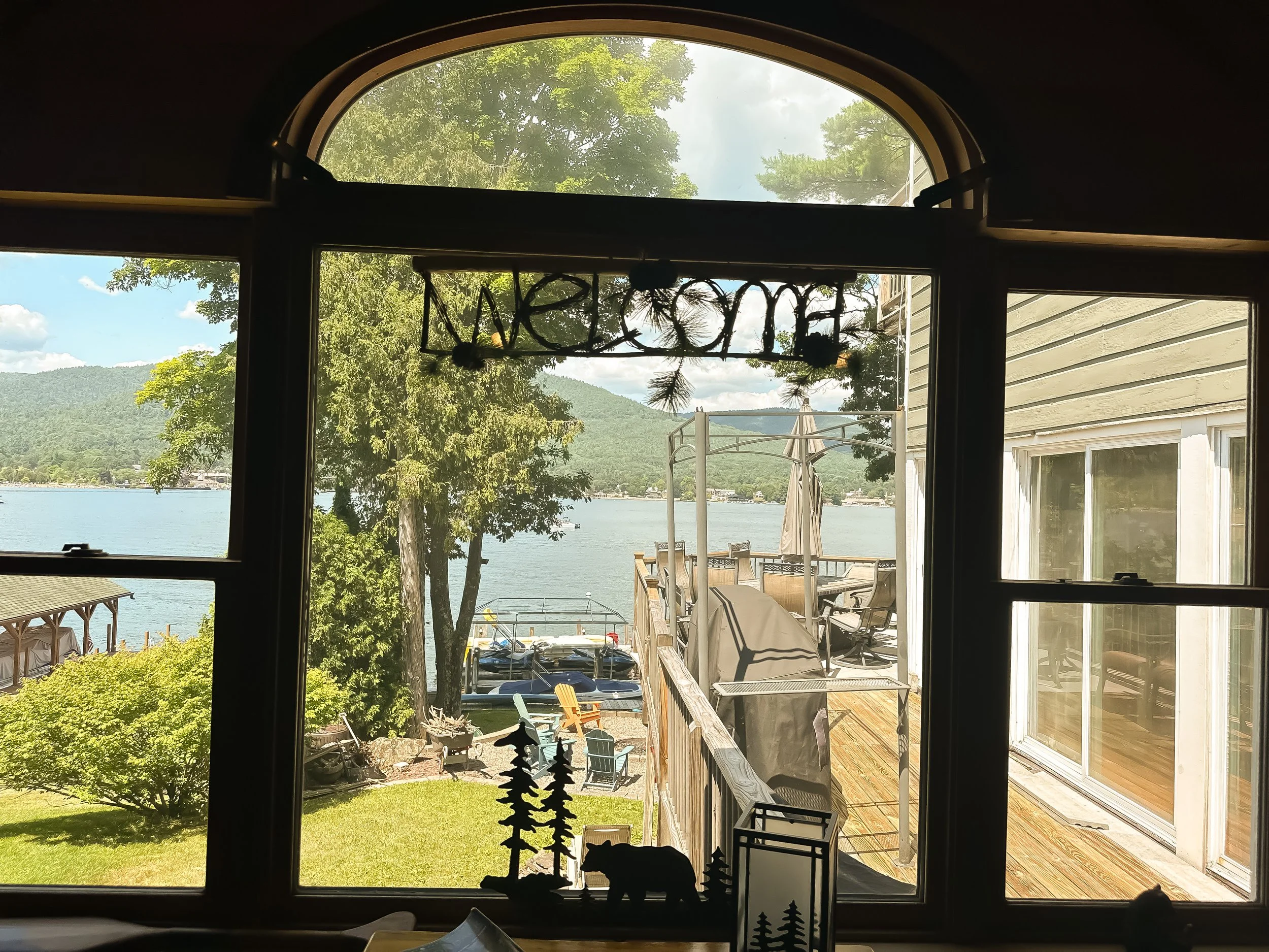 View through a set of glass doors looking out onto a lakeside landscape with trees, houses, and mountains in the background. There are outdoor chairs and a patio umbrella outside, along with some decor items and a dog silhouette in the foreground.