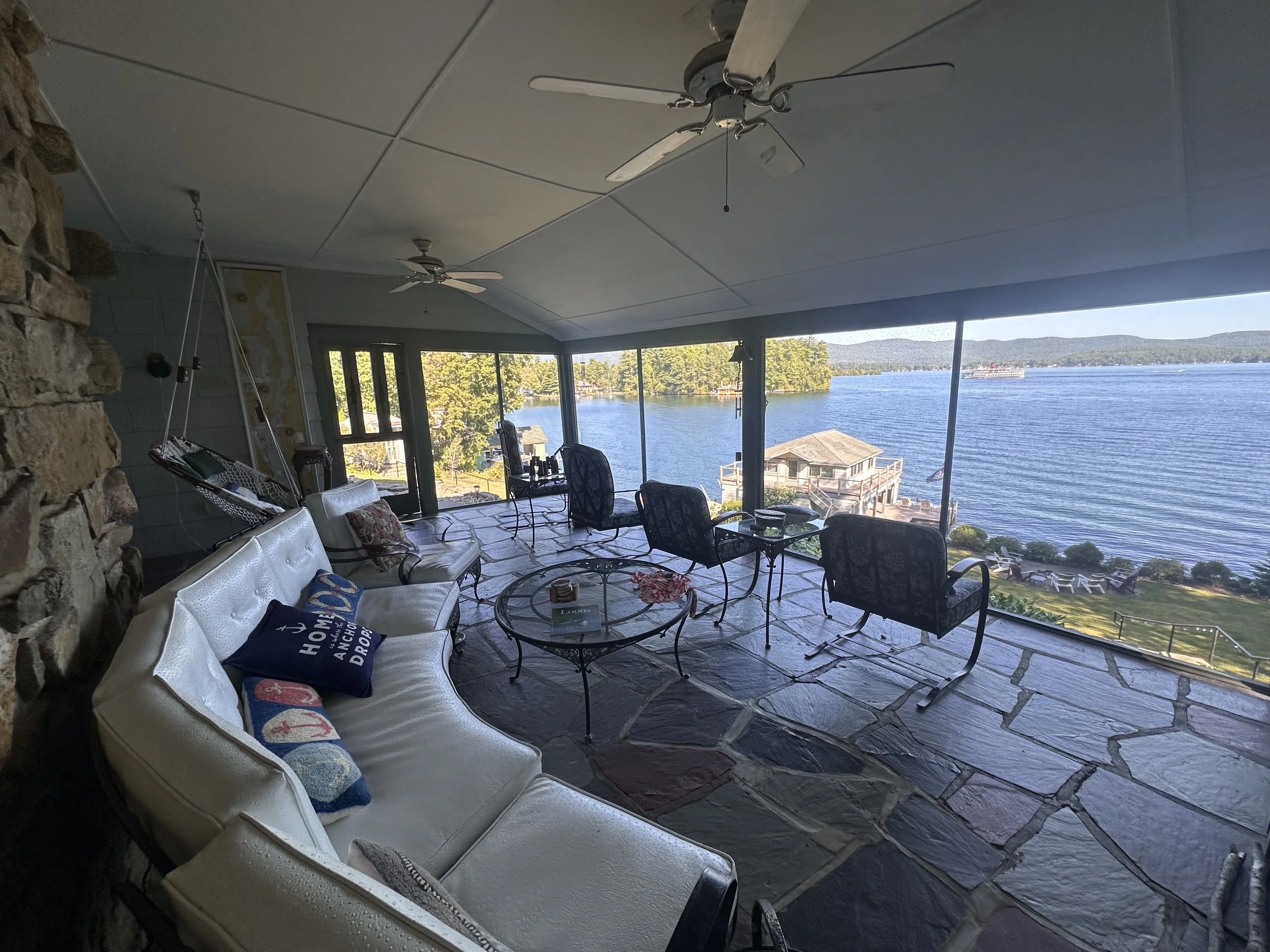 Indoor porch area with stone flooring, white ceiling fans, and view of a large lake through glass windows. There are chairs, a white sofa with throw pillows, and small tables, with outdoor scenery including trees and a dock over the water.