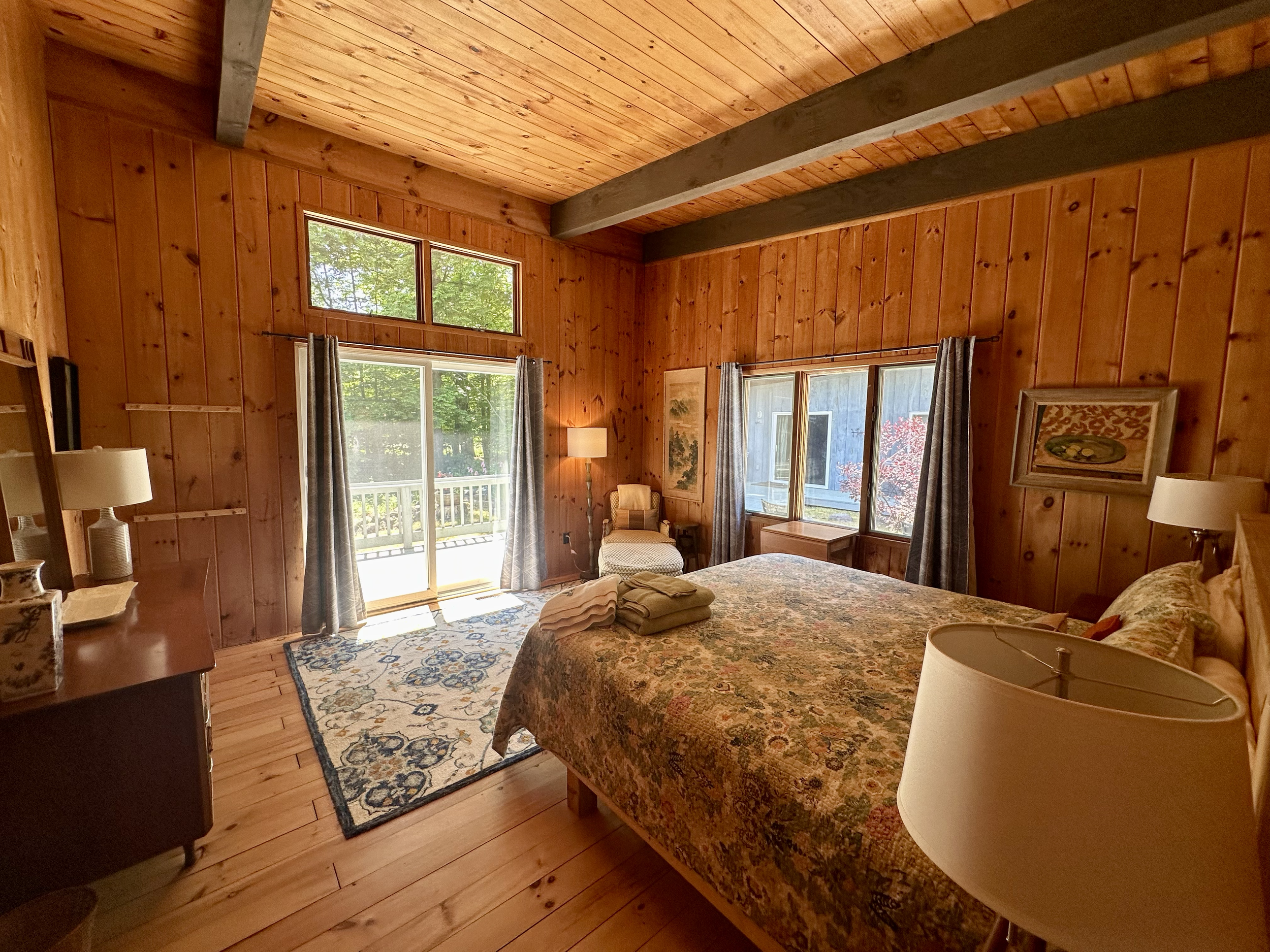 A cozy bedroom with wooden walls, a large bed with a floral quilt, side tables with lamps, a chair by the window, and a sliding glass door leading to a balcony.
