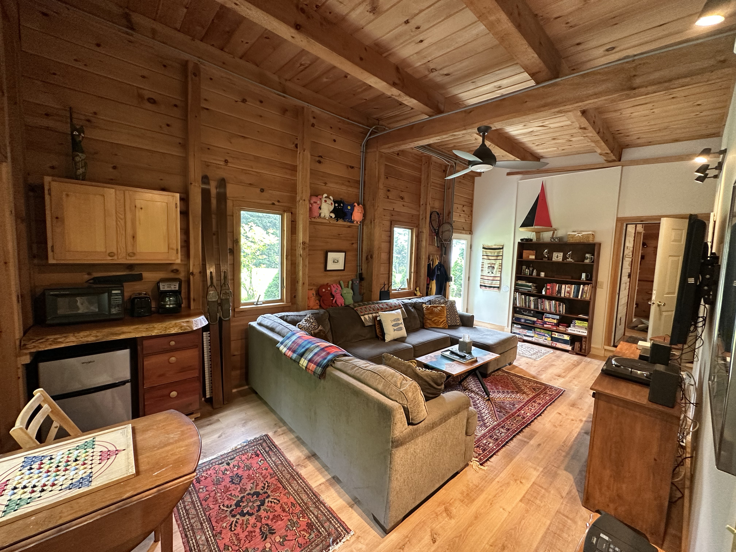 Cozy living room with wood-paneled walls and ceiling, a sectional sofa with pillows, a wooden coffee table, a bookshelf filled with books, and a patterned area rug. Natural light comes through multiple windows, and there are decorations including a m