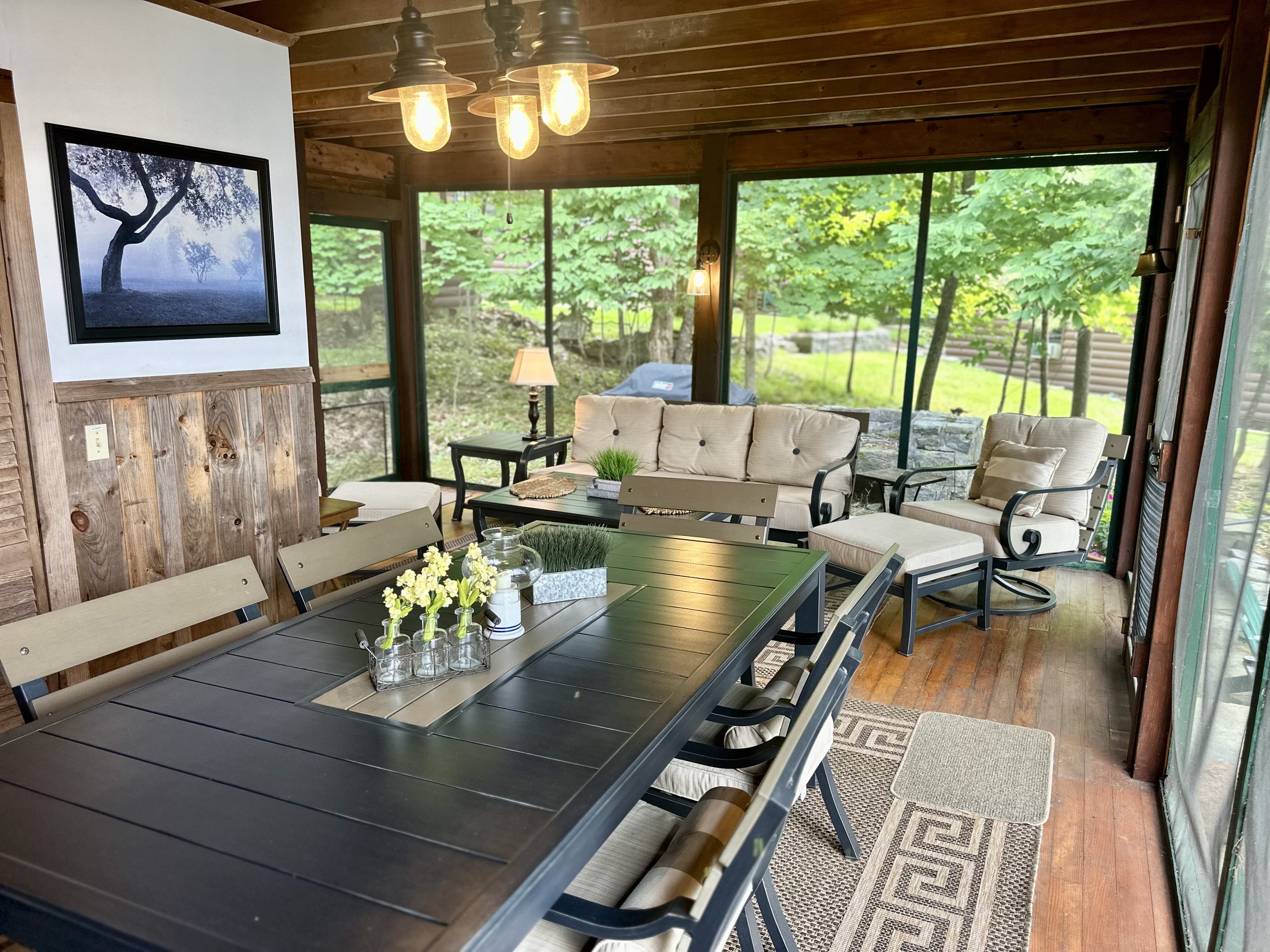 Sunroom with wooden walls, sliding glass windows revealing a green outdoor area, black dining table with chairs, cream-colored cushioned sofa and armchair, side table with a lamp, decorative plants and flowers on the table, framed black and white tre