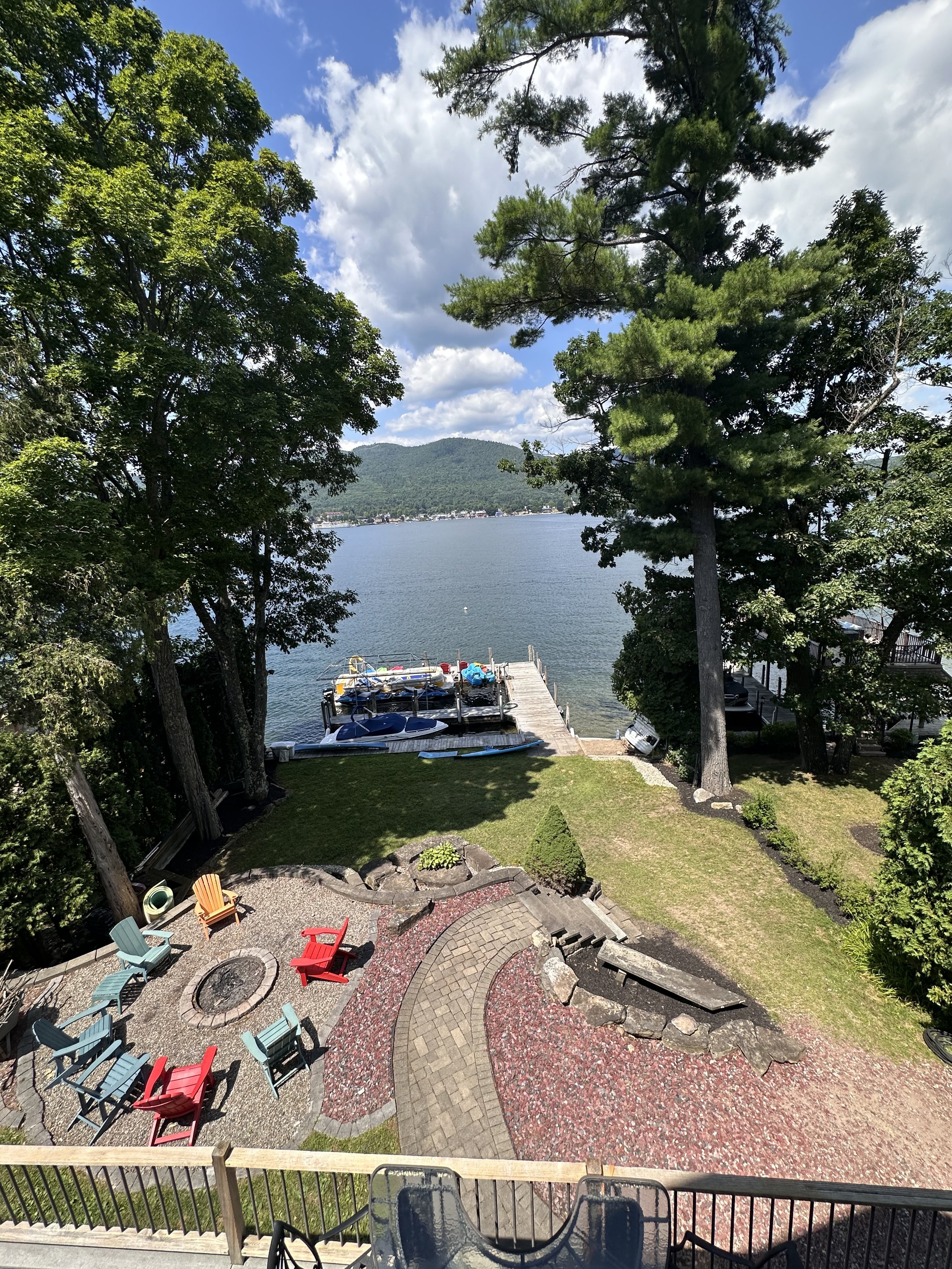 A lakeside backyard scene with trees, a dock with boats, and a mountain in the background. There is a patio with colorful chairs and a fire pit in the foreground.