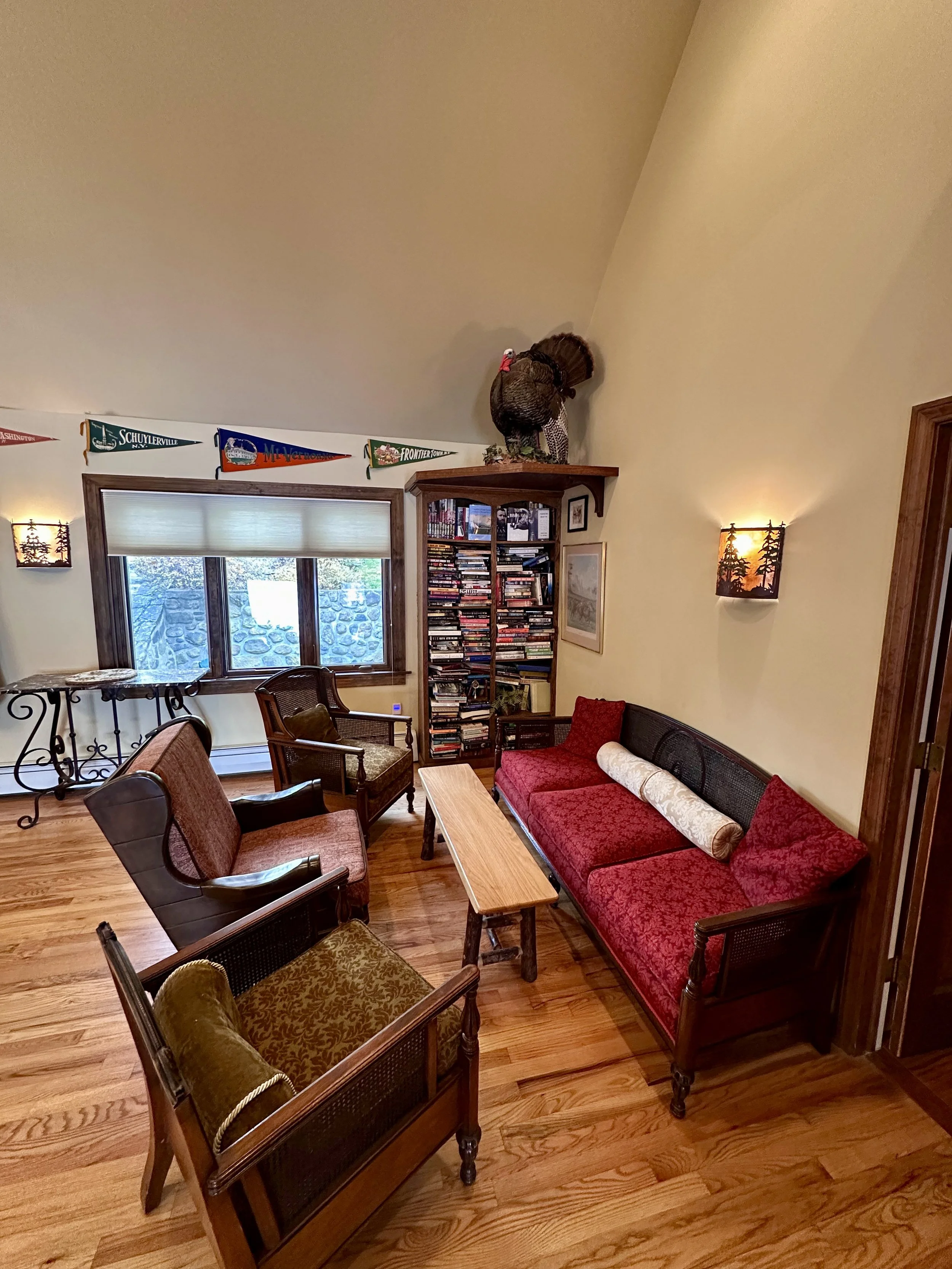 Living room with wooden floor, red sofa with pillows, multiple chairs, window, bookshelf, wall sconces, and a decorative turkey on a shelf.