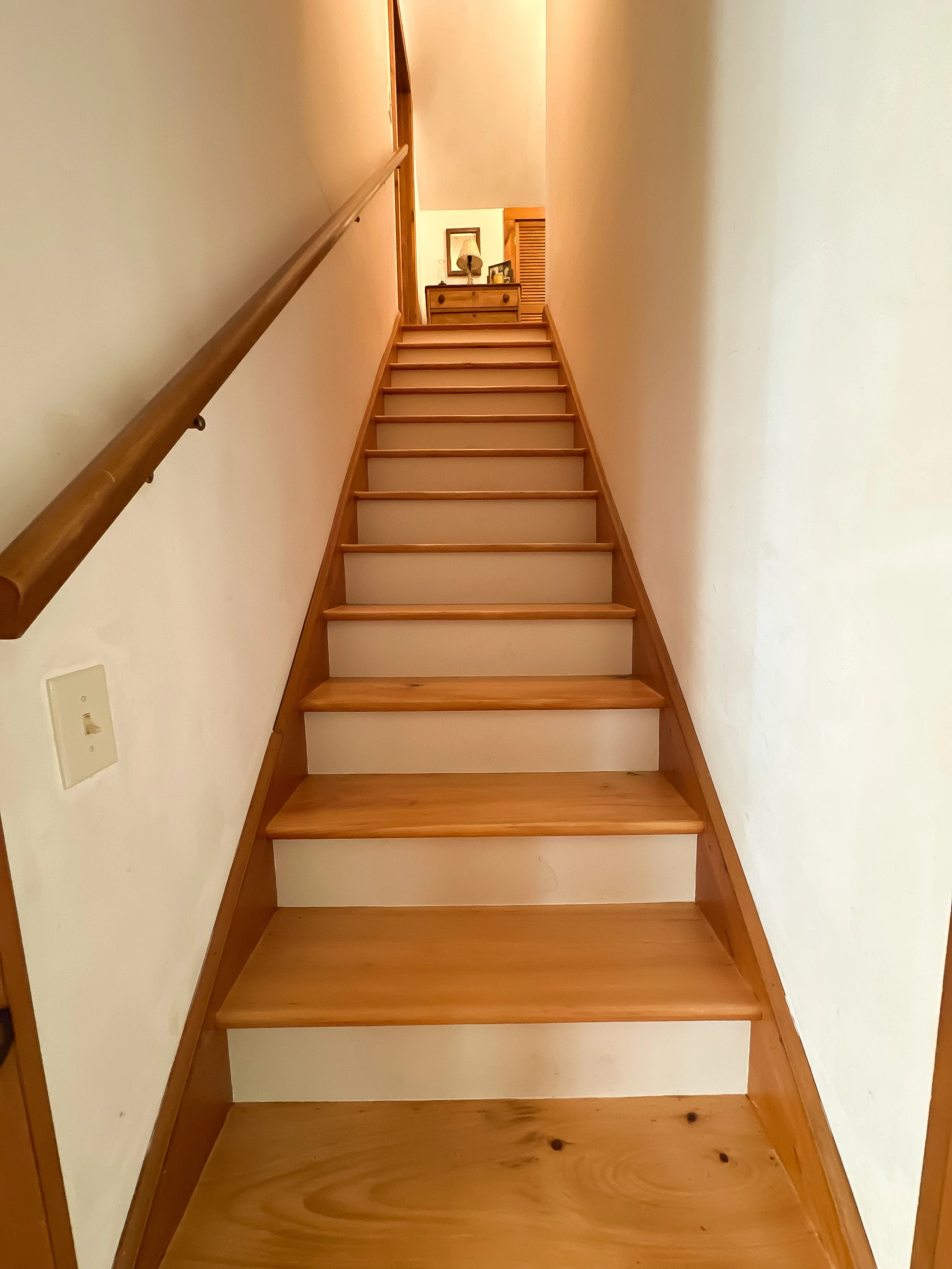 Wooden staircase with a single handrail leading up to a small landing with a dresser and decorative items in a cozy home interior.