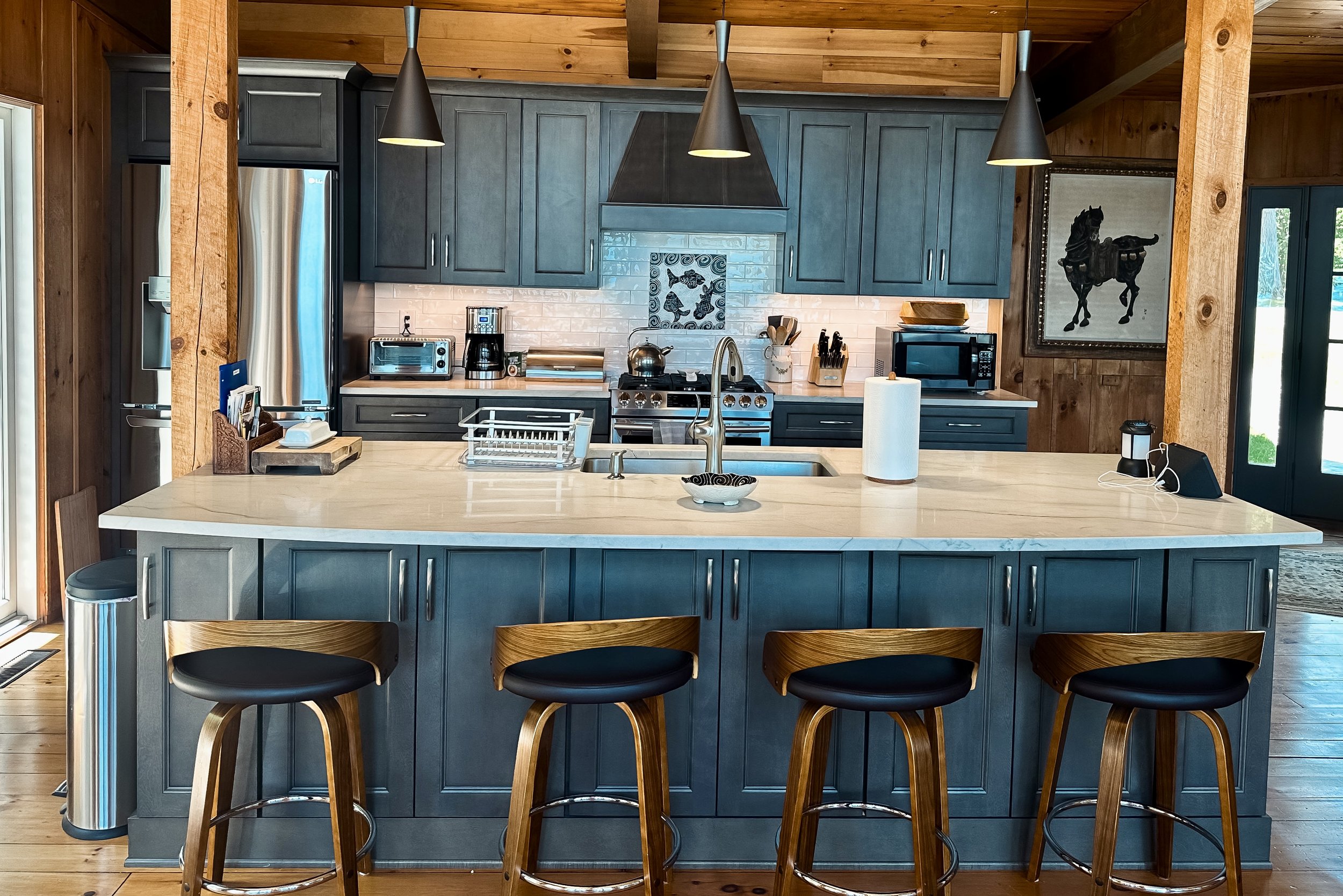 A modern kitchen with blue cabinets, a white countertop island, and wood-paneled walls and ceiling. The island has three wooden bar stools with black seats. The kitchen features stainless steel appliances, including a refrigerator, microwave, and ove
