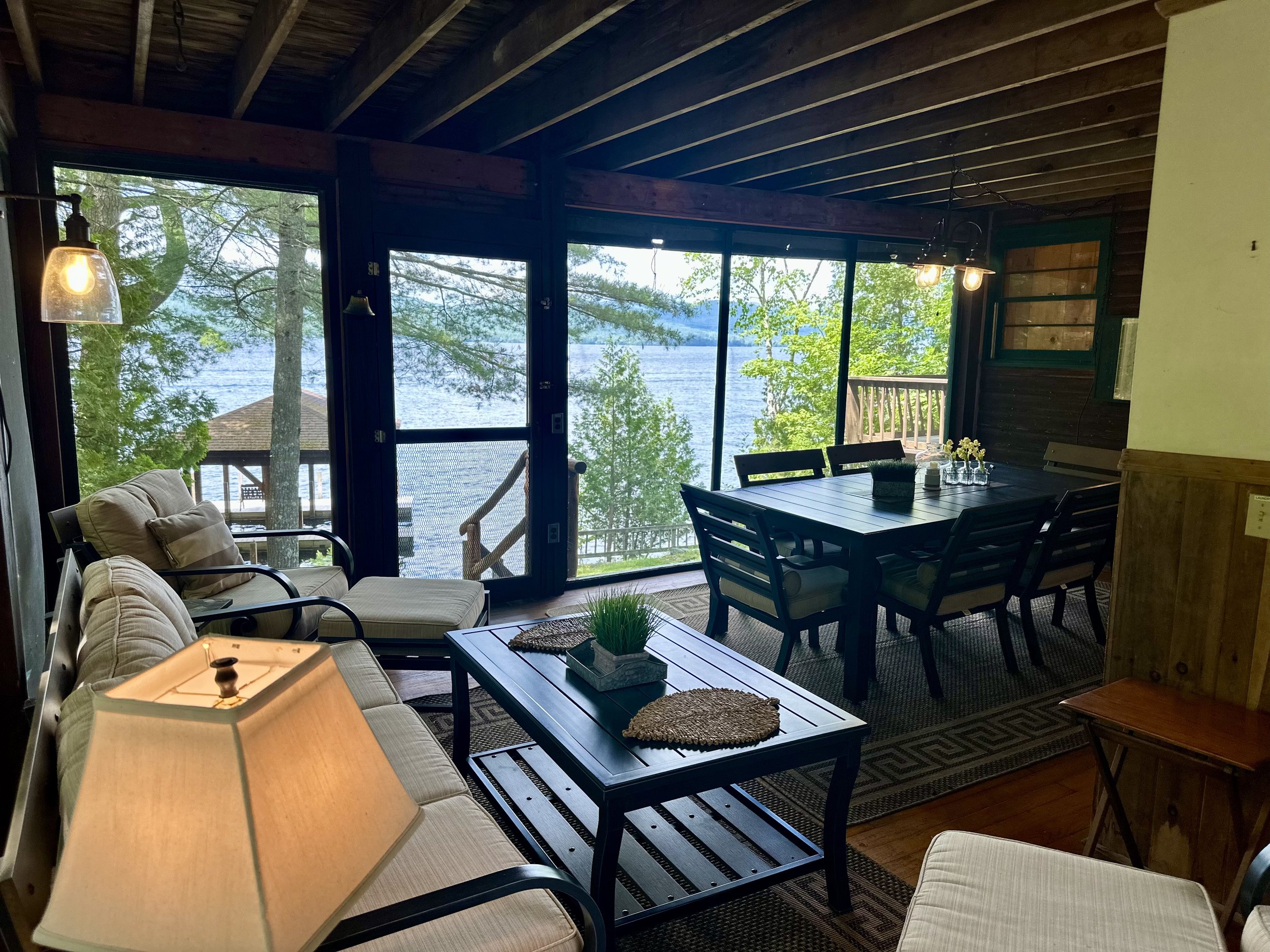 Living room with large windows overlooking a lake, furnished with beige sofas, a wooden dining table with chairs, and a coffee table with decorative items, surrounded by lush green trees.