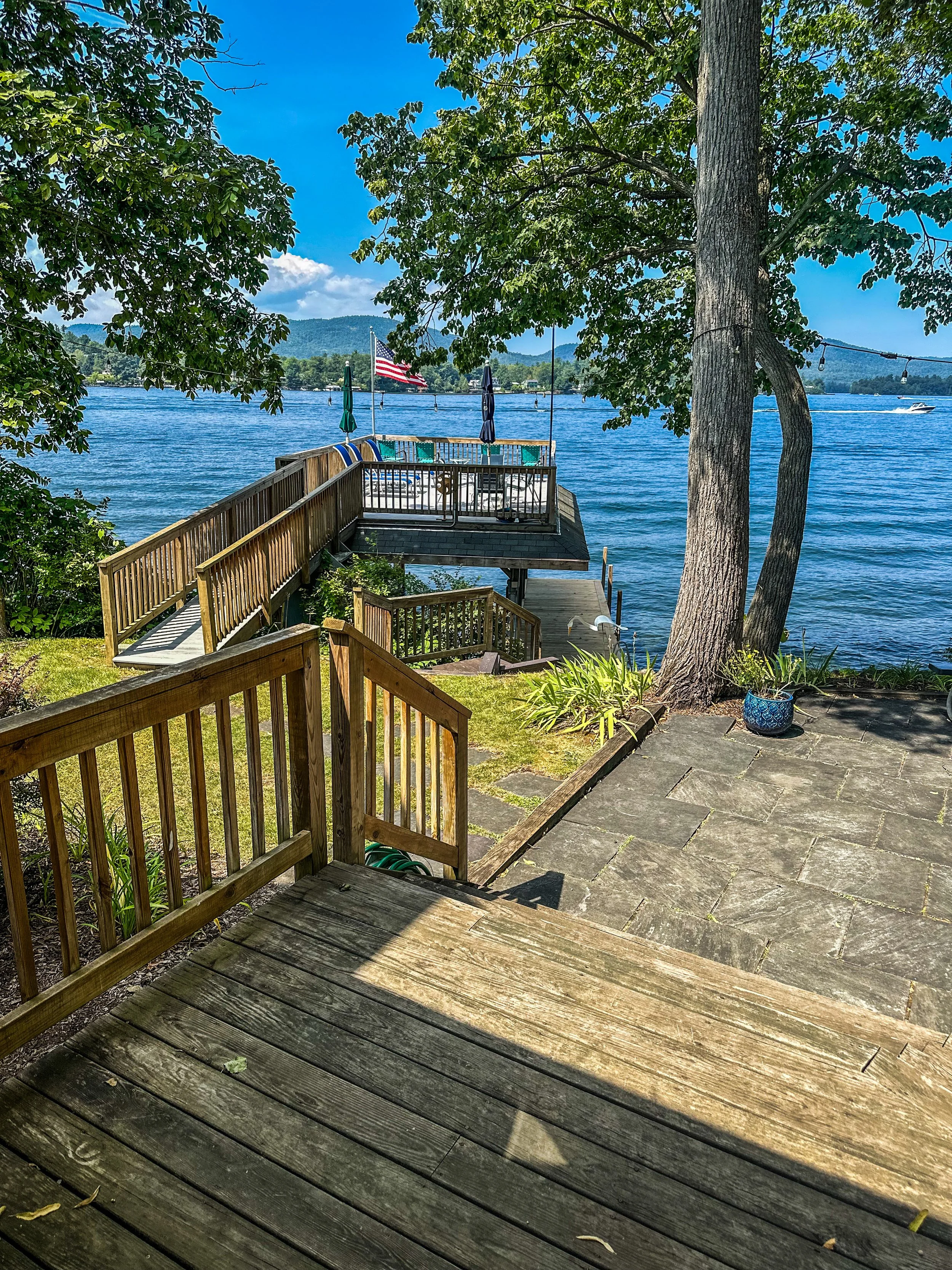 A lakeside view from a wooden deck with steps leading down to a dock, surrounded by trees and greenery, with an American flag, umbrellas, and boats on the water in the background.