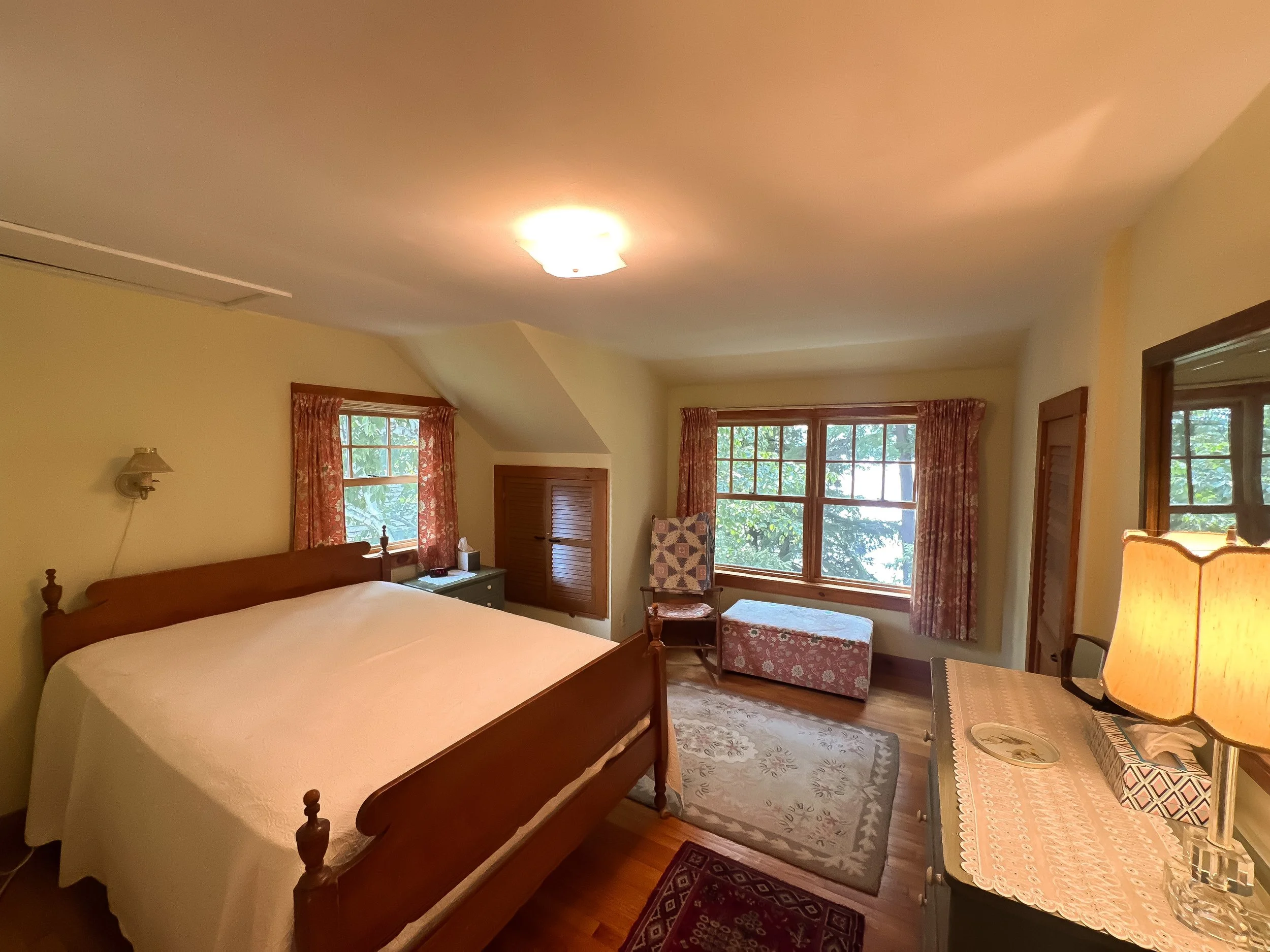 A cozy bedroom with a wooden bed, pink floral curtains, a nightstand, a chair and ottoman by the window, and a dresser with a lamp, illuminated by a ceiling light.
