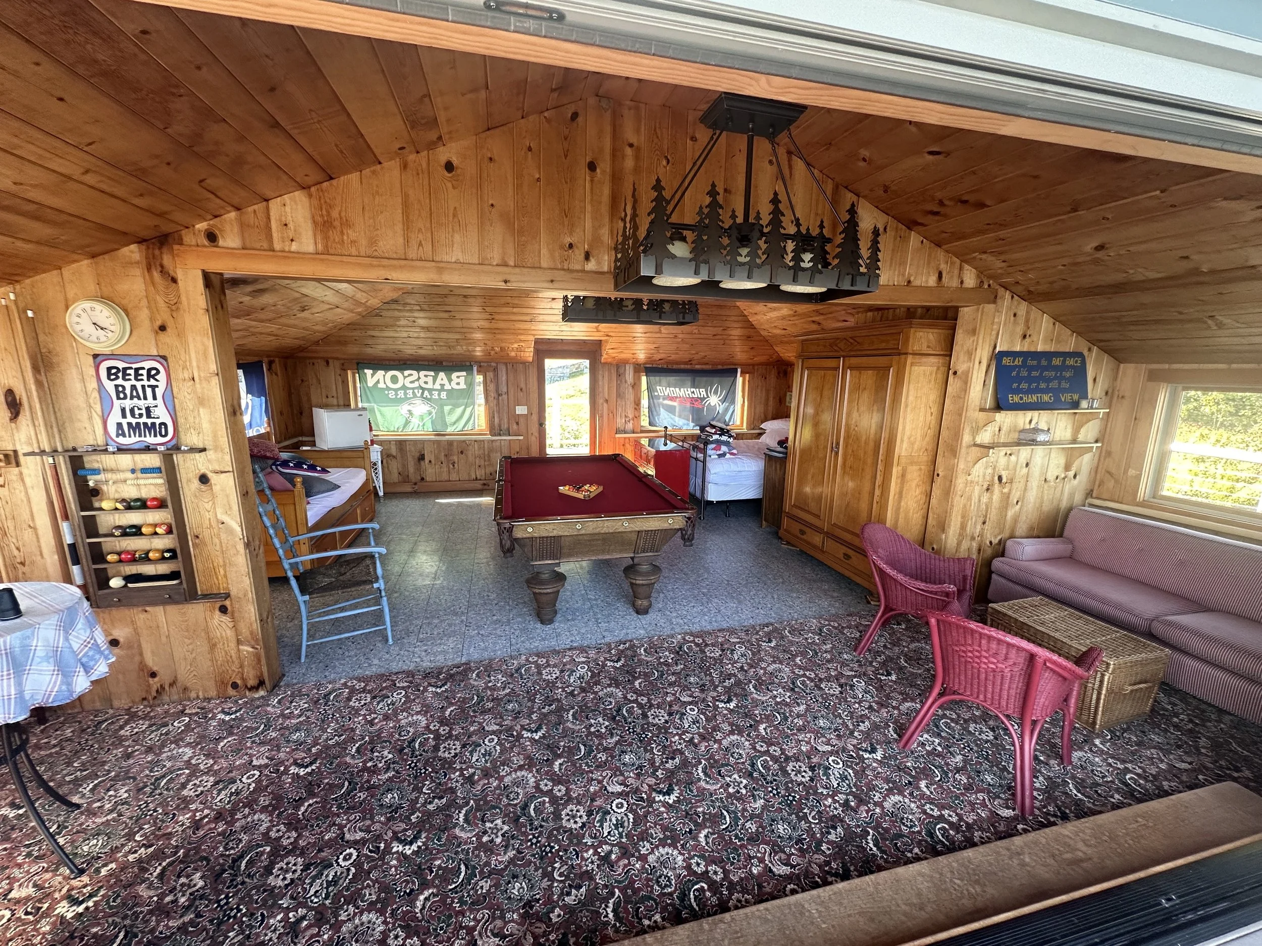 Interior of a rustic wooden cabin with a pool table, sitting area with pink wicker chairs and a pink sofa, two beds, and various decorative signs.