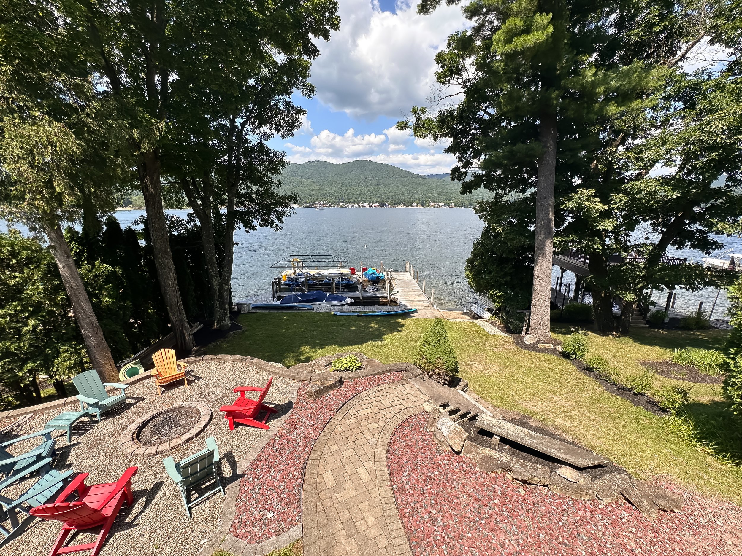 A lakeside backyard with colorful chairs around fire pits, overlooking a lake with boats and dock, framed by trees and mountains in the background.