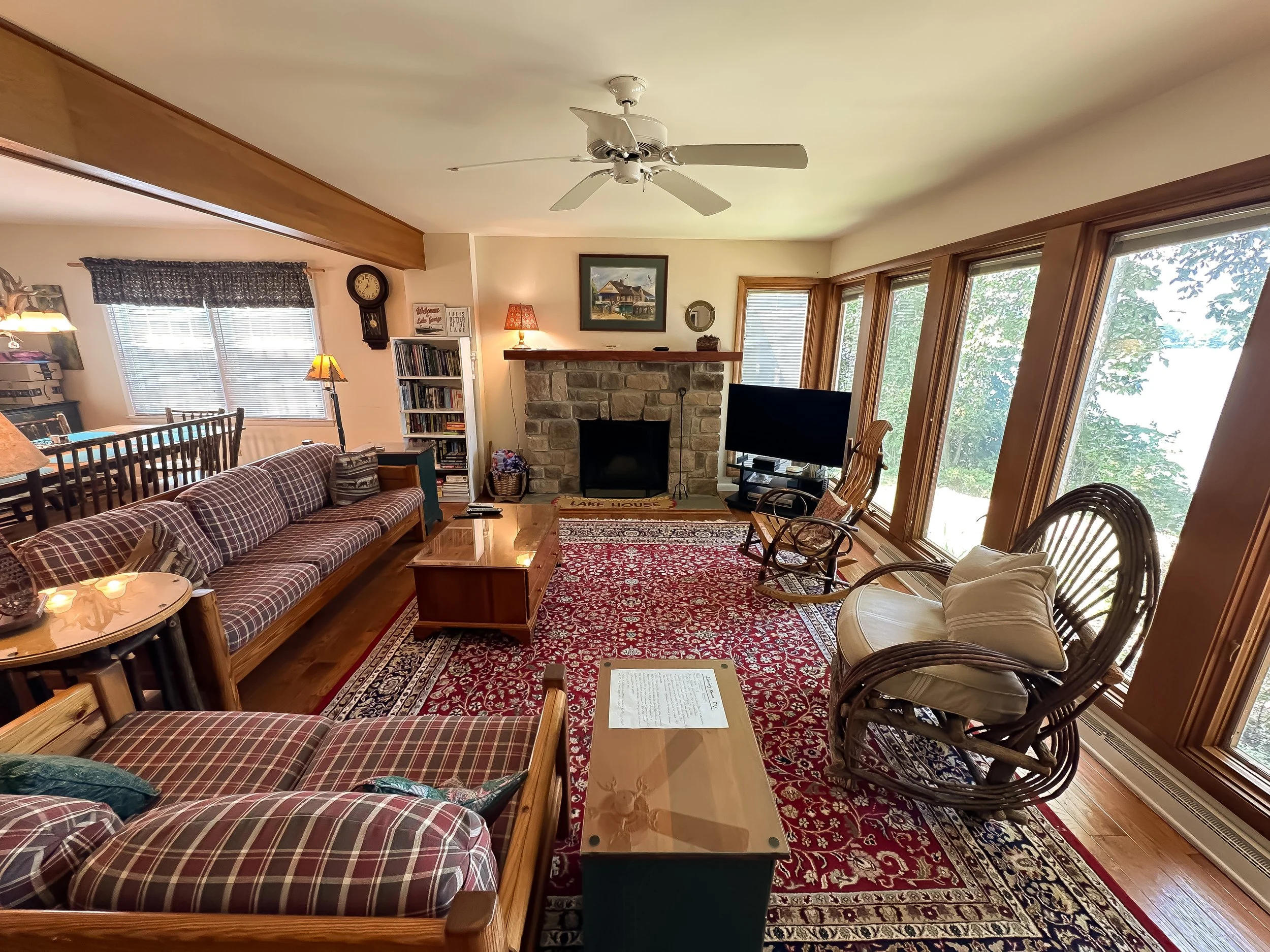 Living room with a plaid fabric sofa, a wooden rocking chair with cushions, a black flat-screen TV, a stone fireplace, and large windows letting in natural light, with a red patterned area rug on the hardwood floor.