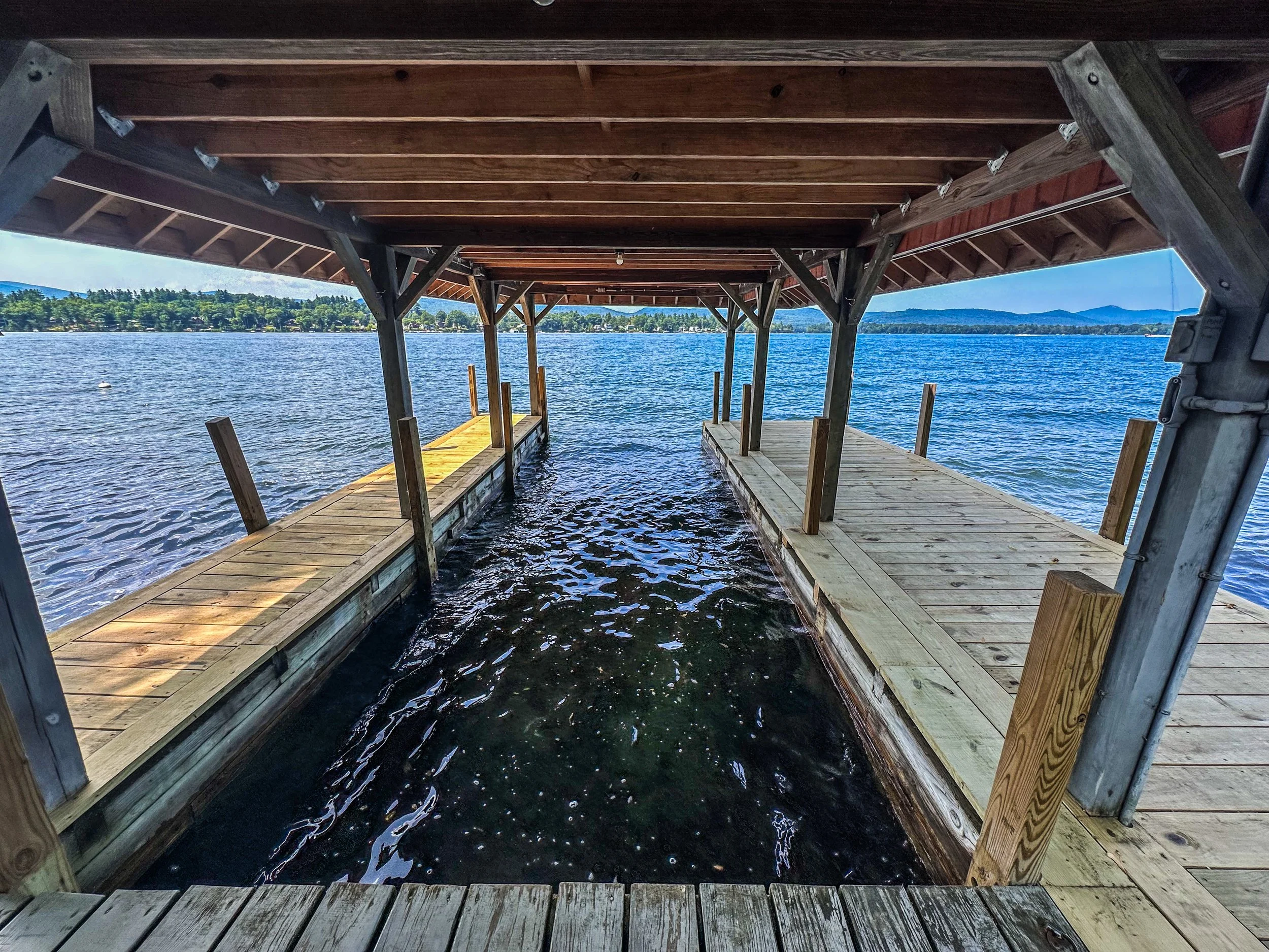 Two wooden boat docks under a covered structure extending into a lake, with calm water, green trees in the background, and mountains in the distance.