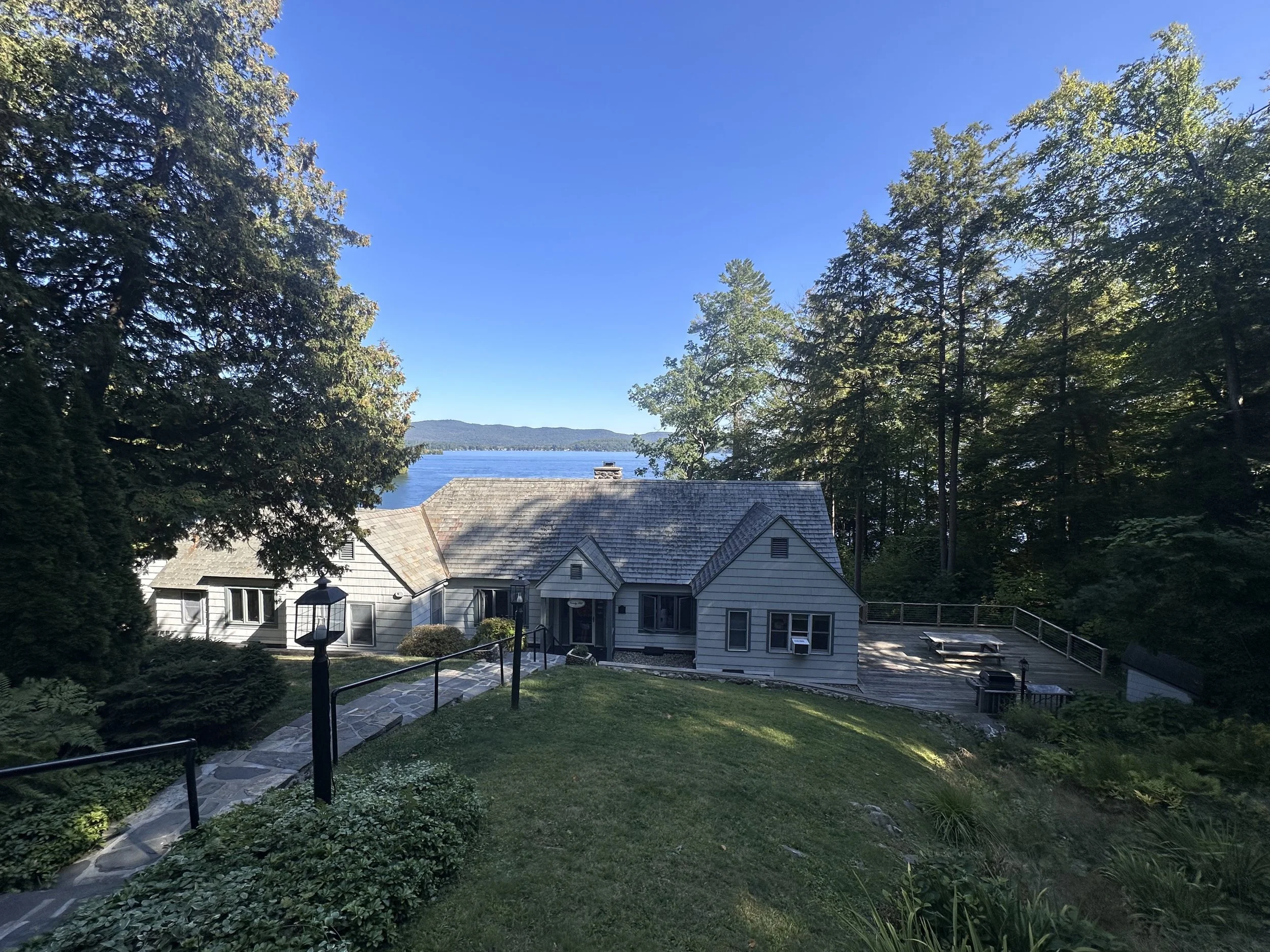 A house by the water with a dock and picnic table, surrounded by trees and greenery on a sunny day.
