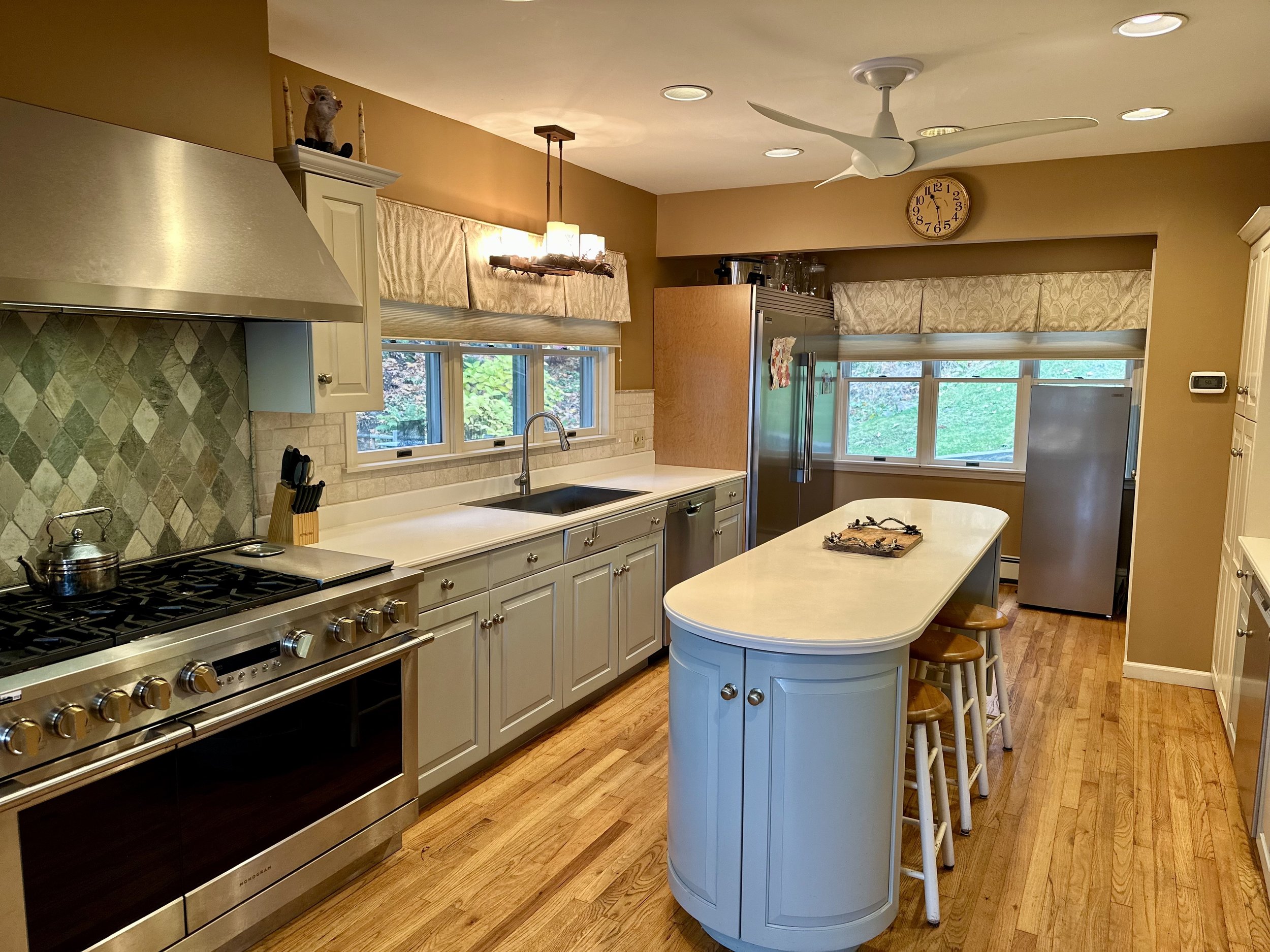 A spacious kitchen with light gray cabinets, a stainless steel stove, a large kitchen island with three stools, and multiple windows with beige valances. The room has hardwood floors, a ceiling fan, and warm-toned walls.