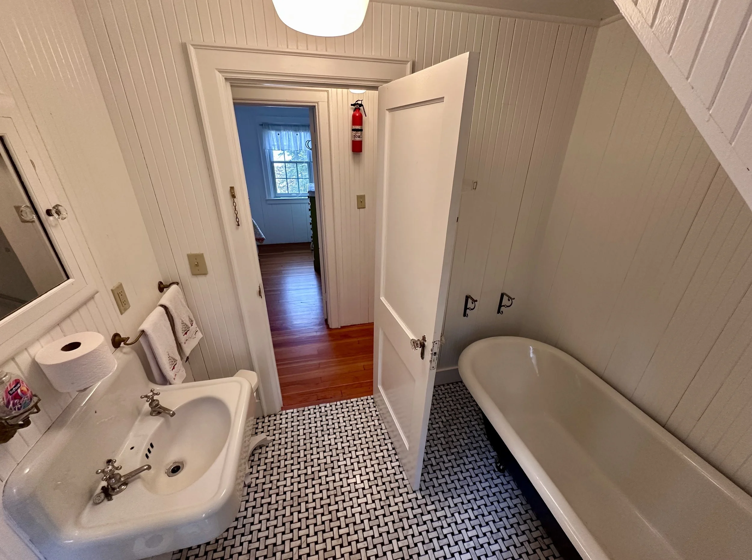 A vintage bathroom with a clawfoot bathtub, a white sink with two silver faucets, a mirror, a hand towel hanging, a toilet paper roll, a fire extinguisher on the wall, and a view into a room with wooden floors and a window.