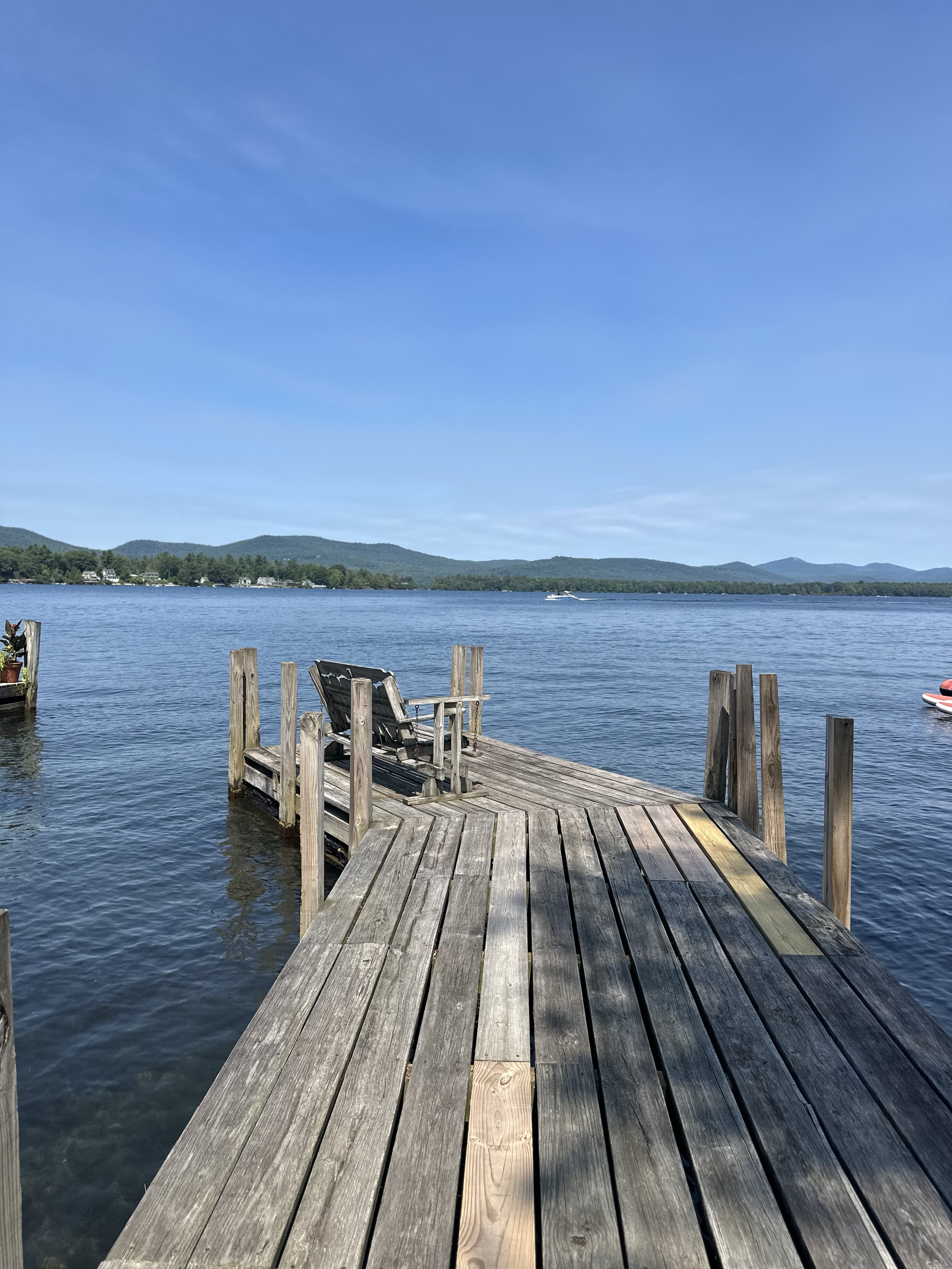 A wooden dock extends into a calm lake with a chair at the end, surrounded by water and distant green hills under a blue sky.