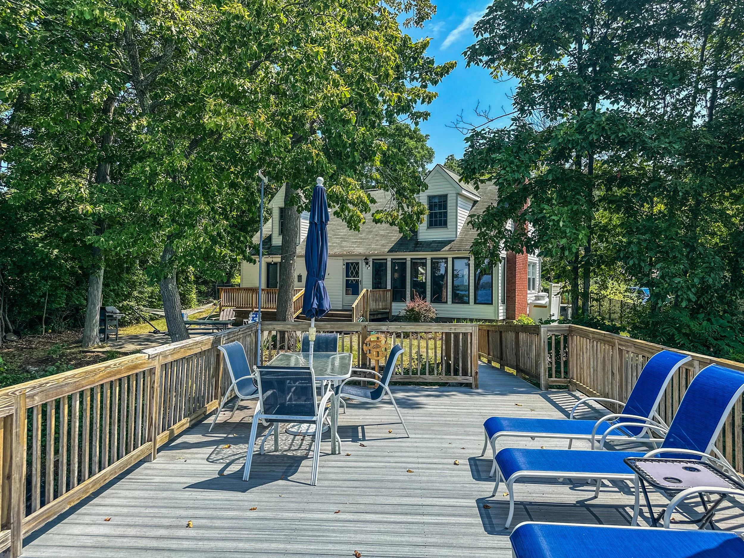 Sunlit wooden deck with outdoor furniture, including a glass table with six chairs and three blue lounge chairs, surrounded by trees and a house in the background.