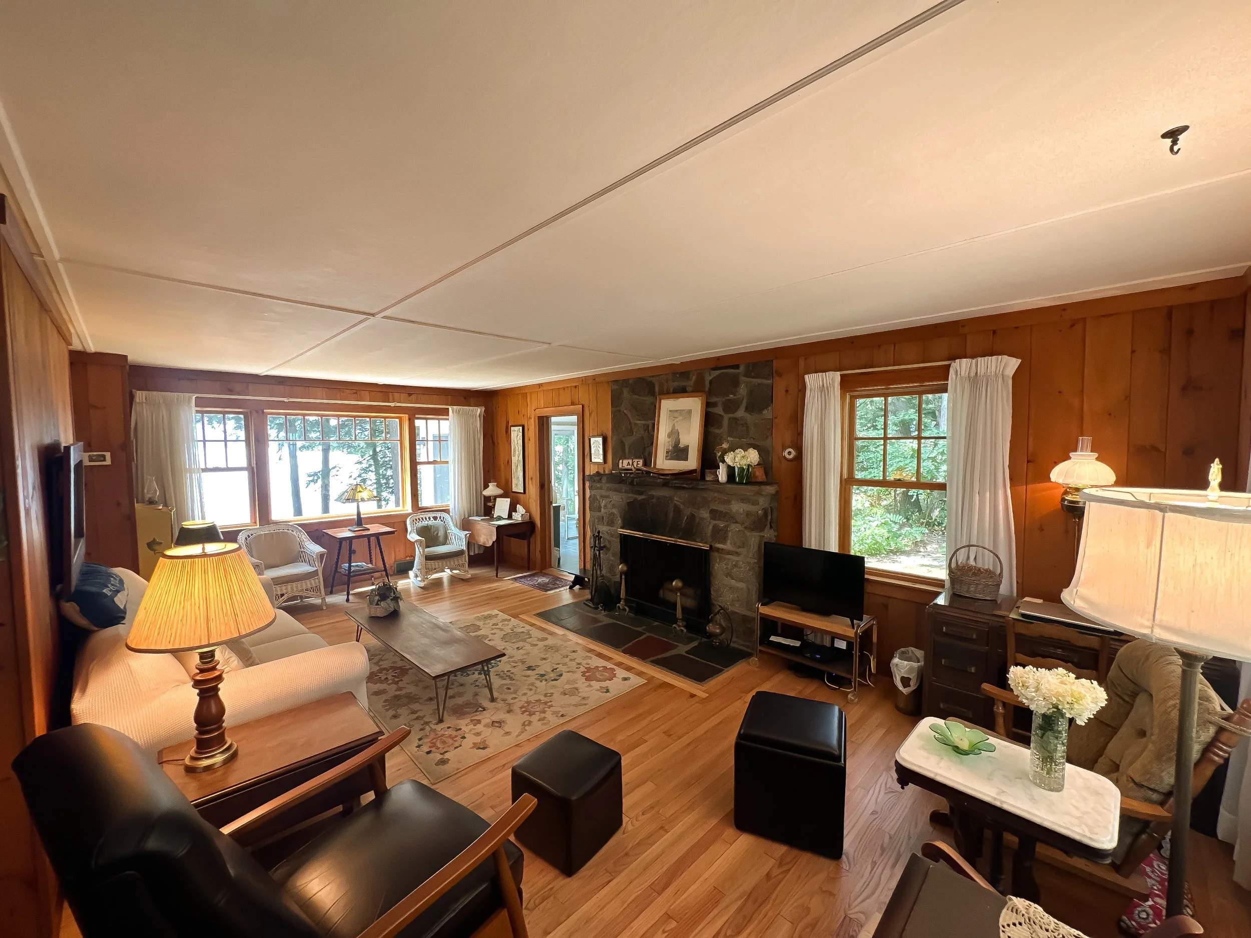 Living room with wood-paneled walls, stone fireplace, multiple windows with white curtains, various armchairs, a sofa, lamps, television, and a wooden coffee table on hardwood floors.