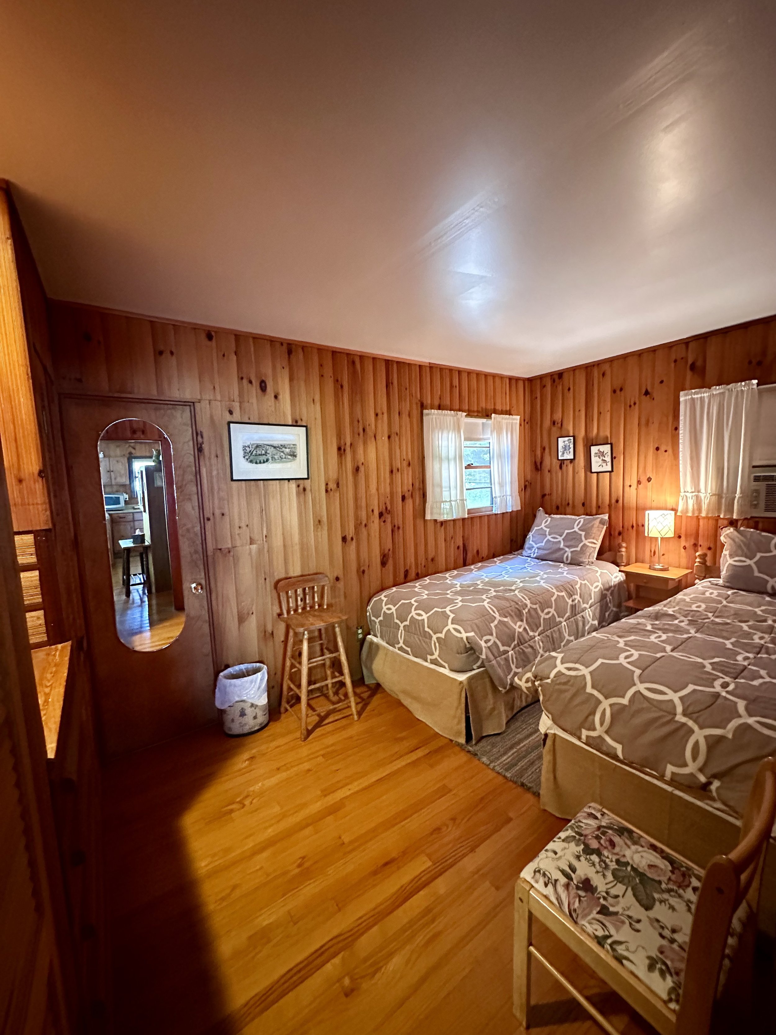 A cozy bedroom with wood-paneled walls, two single beds with patterned bedding, small wooden furniture, a window with curtains, and a wooden chair with floral upholstery.