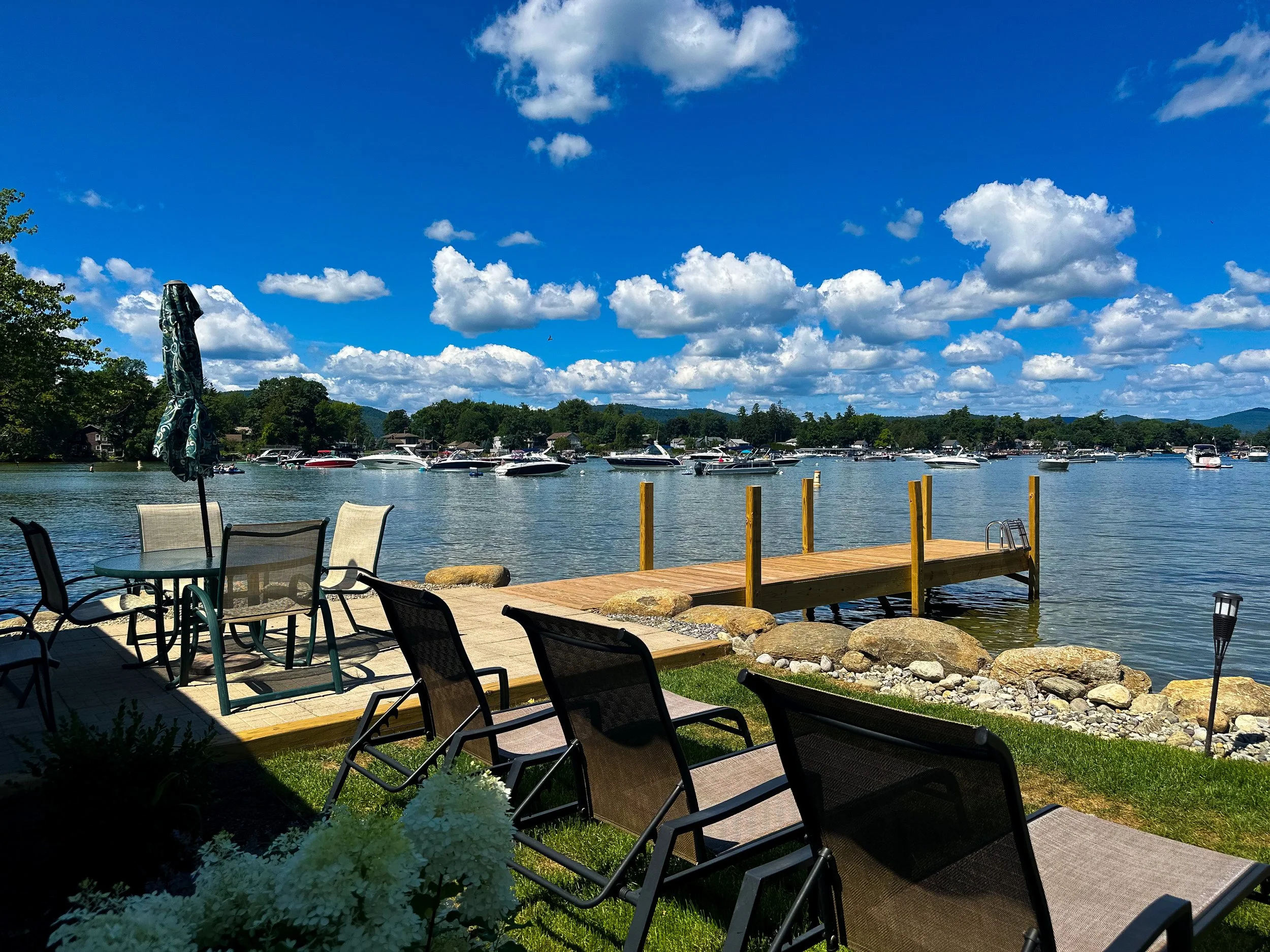 Scene of a lakeside view with boats docked, a wooden pier, outdoor chairs and tables, umbrellas, and a partly cloudy blue sky.