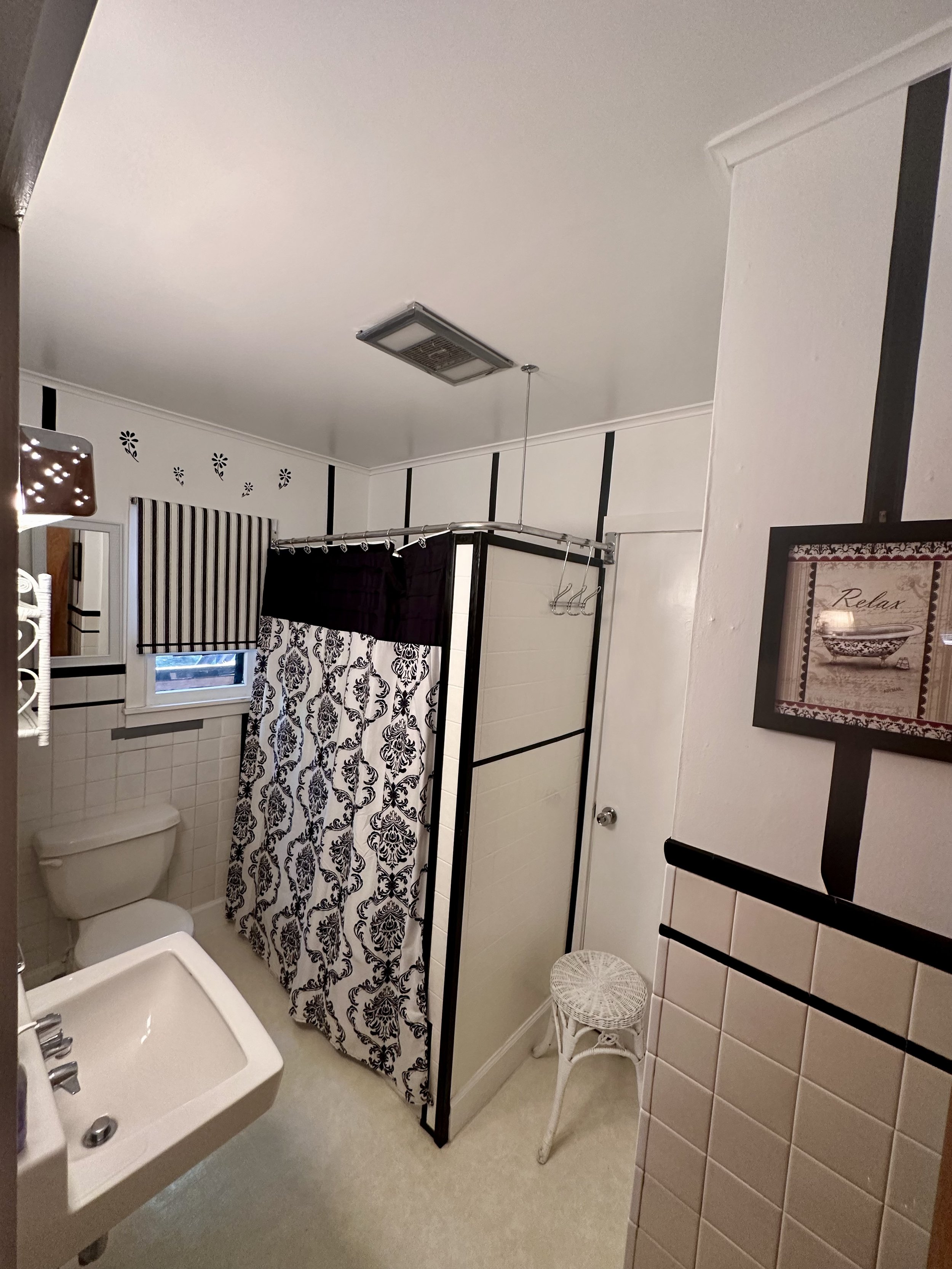 Small bathroom with a white toilet, beige sink, and black-and-white patterned shower curtain. A window with striped and solid black curtains, a white wicker stool, and black vertical accents on the white walls. Decorative wall art with a relaxing the
