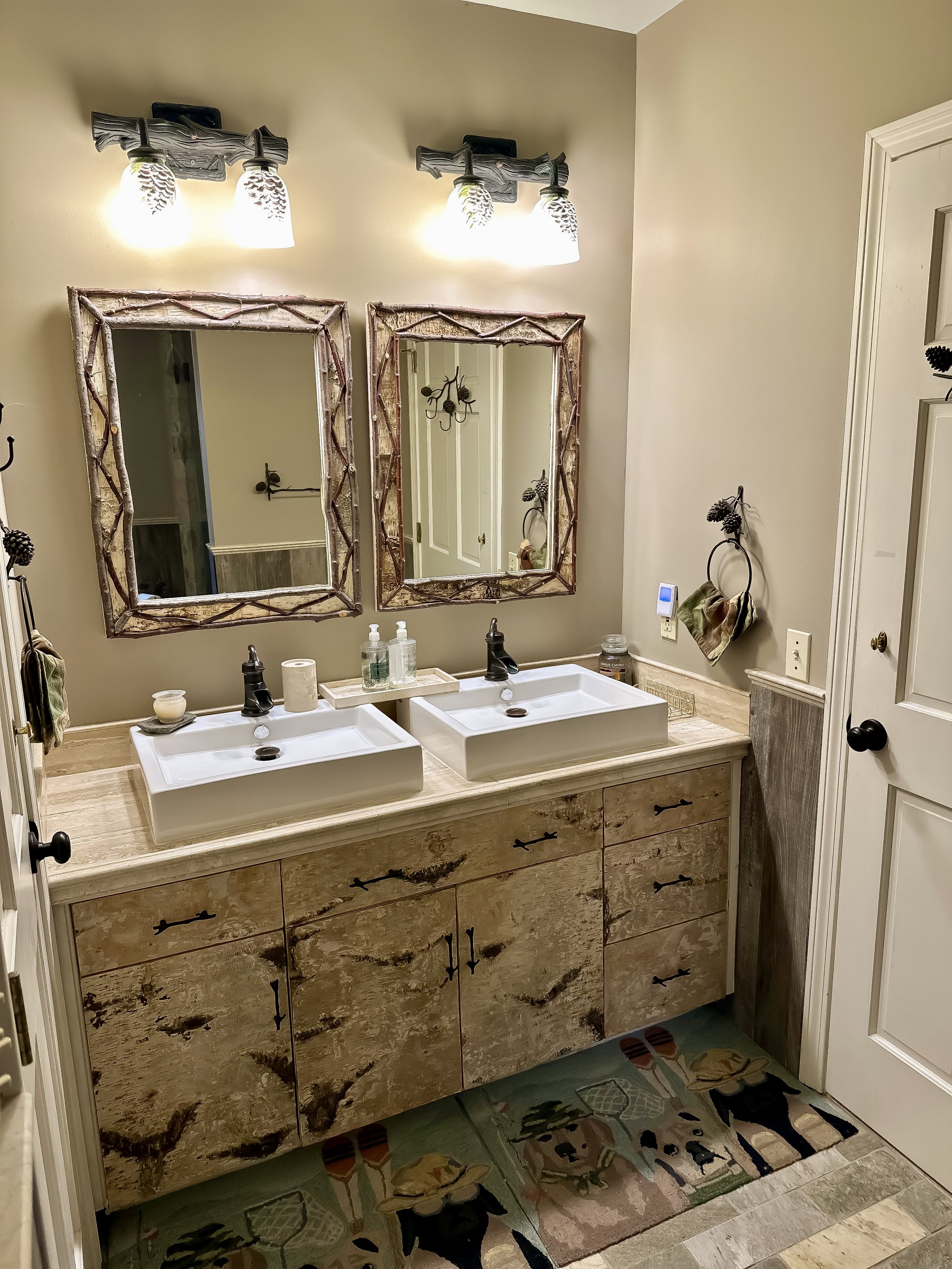 Bathroom with a double vanity, two white sinks, two mirrors with rustic frames, wood cabinetry, and two light fixtures with three bulbs each.