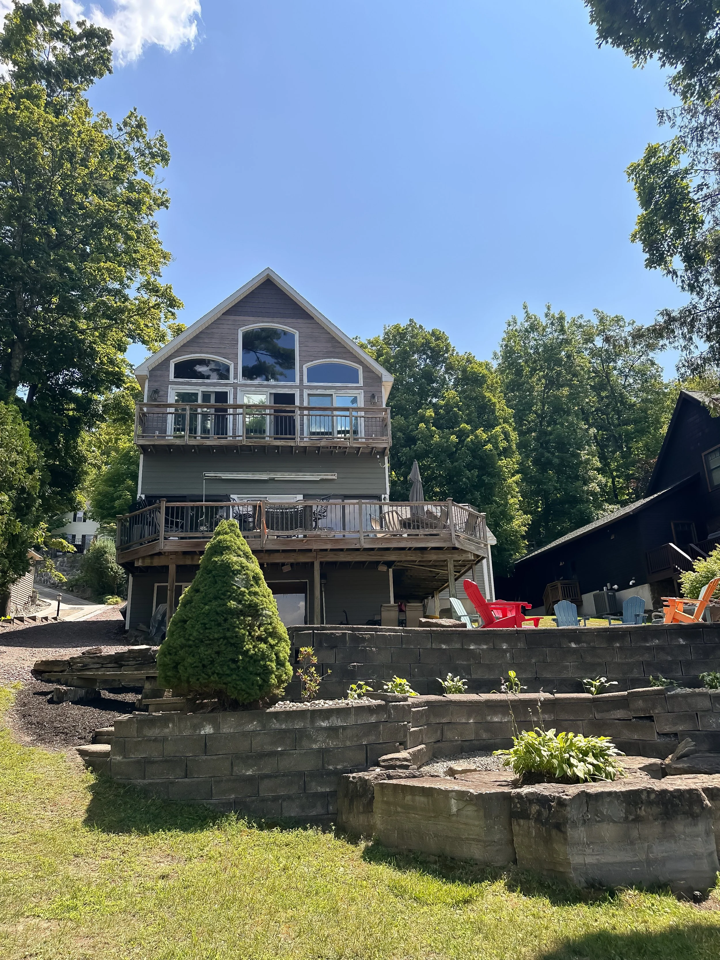 A multi-story house with large windows and a wooden balcony, situated in a lush yard with a small fountain, paved walkways, and colorful outdoor chairs, surrounded by tall green trees on a sunny day.