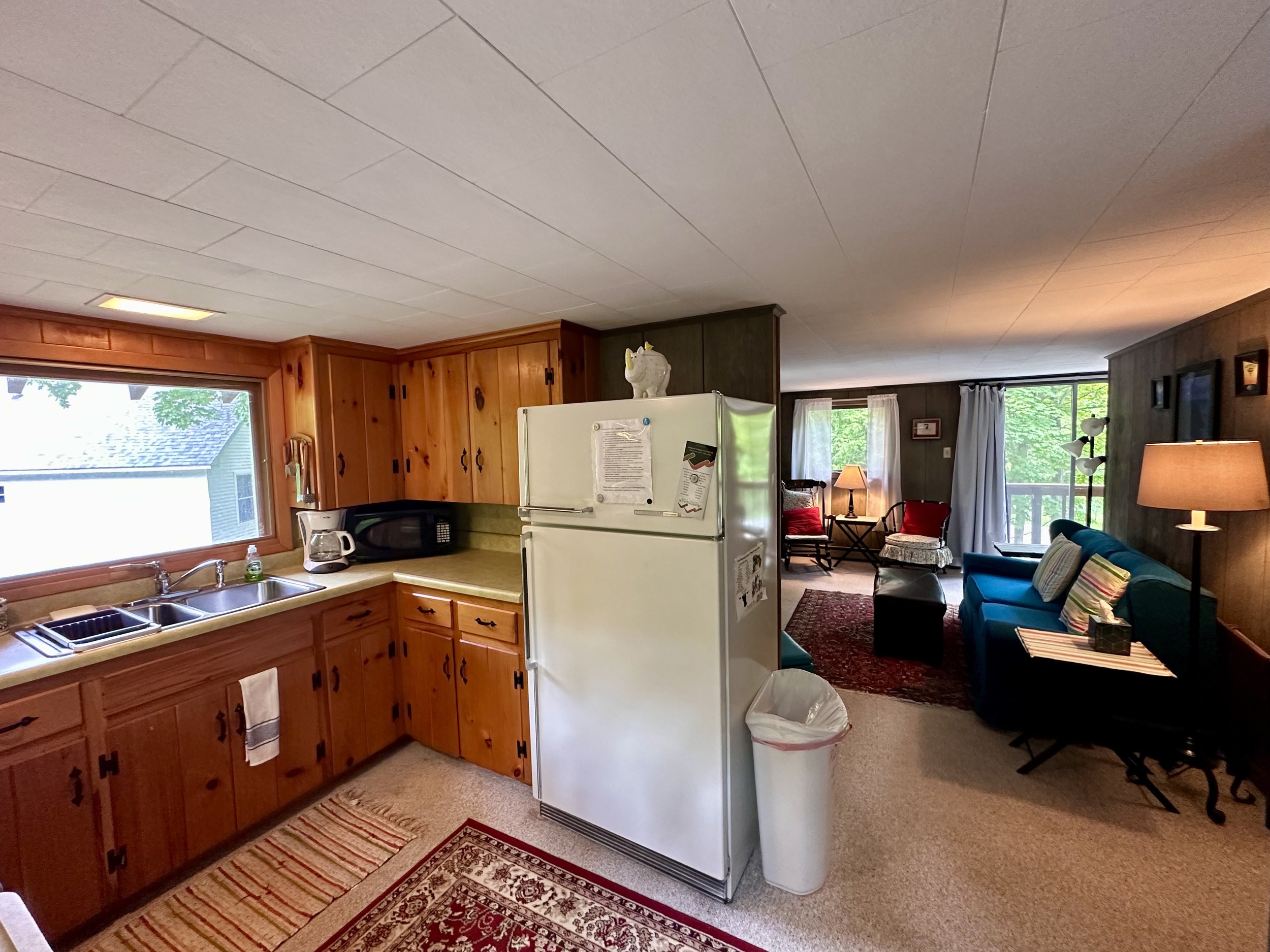 Kitchen with wooden cabinets, window, white refrigerator, microwave, coffee maker, and living room with blue sofa, armchairs, lamps, and large windows in a cozy home.